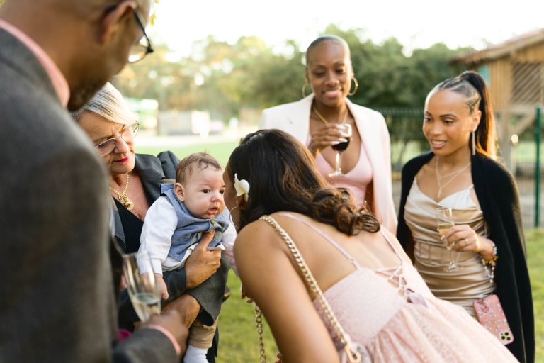 Un groupe de six adultes, habillés en tenue de soirée, se rassemblent à l'extérieur autour d'une personne tenant un bébé. Le bébé porte une tenue bleue et regarde une femme qui se penche vers lui. D'autres sourient et tiennent des verres, peut-être lors d'une célébration de mariage Audenge dans un jardin.