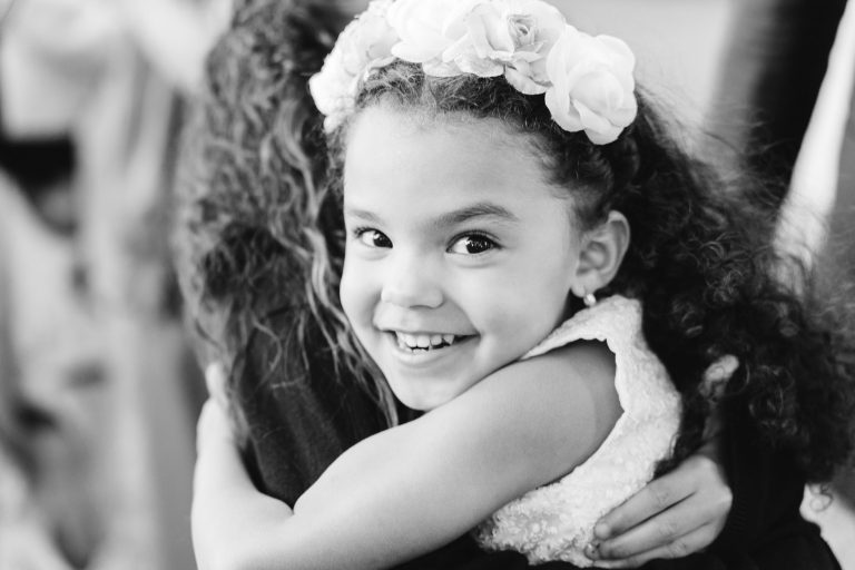 Image en noir et blanc d'une jeune fille aux cheveux bouclés, portant un bandeau fleuri et une robe en dentelle. Elle sourit joyeusement tout en embrassant quelqu'un qui tourne le dos à la caméra lors d'un Mariage Audenge. L'arrière-plan est flou, attirant l'attention sur son visage expressif et heureux.