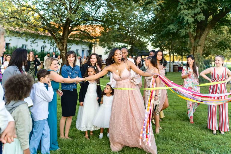 Une femme vêtue d'une robe rose scintillante coupe un ruban avec de gros ciseaux lors d'un rassemblement en plein air, célébrant le mariage Audenge. Elle est entourée d'hommes, de femmes et d'enfants souriants, certains habillés de façon formelle. Des rubans colorés sont attachés au ruban principal. Des arbres et un bâtiment sont en arrière-plan lors d'une journée ensoleillée.