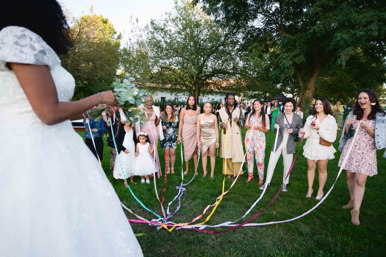 Une mariée en robe blanche tient un bouquet et des rubans colorés tandis qu'un groupe de femmes et d'enfants se tient dans une zone herbeuse, chacun tenant un ruban. Des arbres et un bâtiment sont visibles en arrière-plan. Les femmes, participantes au Mariage Audenge, sont habillées de diverses tenues, profitant de ce moment de célébration.