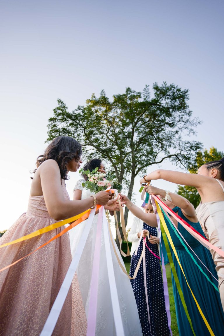 Une mariée en robe blanche et tenant un bouquet est entourée de demoiselles d'honneur vêtues de robes colorées, capturant l'essence de Mariage Audenge. Elles sont à l'extérieur, profitant d'une journée ensoleillée. Chaque demoiselle d'honneur tient un ruban de différentes couleurs, créant une atmosphère festive alors qu'elles se dirigent vers la mariée, qui sourit joyeusement. De grands arbres sont visibles en arrière-plan.