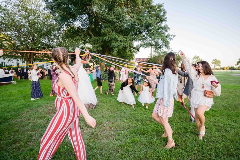 Une fête en plein air animée au cours de laquelle des personnes, dont des enfants vêtus de blanc, s'adonnent au tissage de rubans colorés autour d'un mât de cocagne. L'événement, qui rappelle un mariage Audenge, se déroule sur un terrain herbeux avec de grands arbres fournissant de l'ombre. Les participants portent des tenues diverses tandis que d'autres regardent et apprécient en arrière-plan.