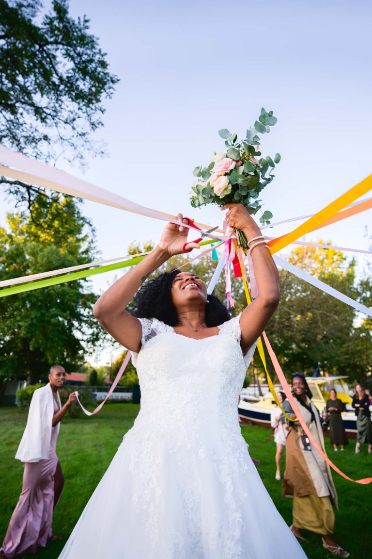 Une mariée en robe de mariée en dentelle blanche lève son bouquet, entrelaçant des rubans colorés avec ses amis sur une pelouse verte luxuriante sous un ciel clair. Les arbres et les invités, certains portant plus de rubans, rehaussent l'atmosphère festive de célébration en plein air de ce Mariage Audenge enchanteur.