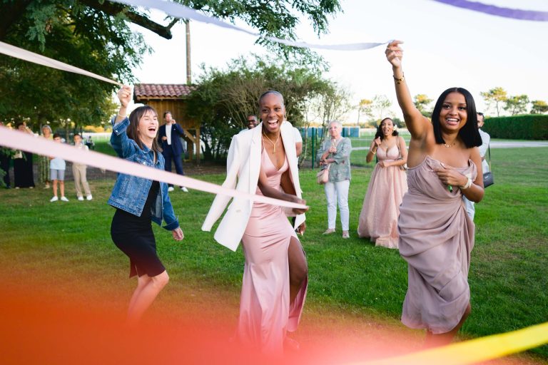 Un groupe de personnes profite d'un événement en plein air ensoleillé. Trois femmes au premier plan rient et jouent avec des rubans colorés, portant des robes élégantes ; l'une en blazer blanc, une autre en robe mauve et la dernière en robe bustier. D'autres personnes présentes au Mariage Audenge regardent et sourient.