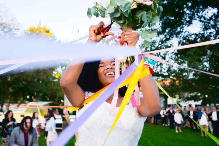 Lors d'un mariage Audenge, une mariée vêtue d'une robe blanche coupe des rubans colorés avec une paire de ciseaux rouges. Elle sourit et la scène semble se dérouler en extérieur dans un parc avec des arbres en arrière-plan. Une foule de personnes se tient à l'arrière-plan, certaines floues, et regardent la cérémonie.