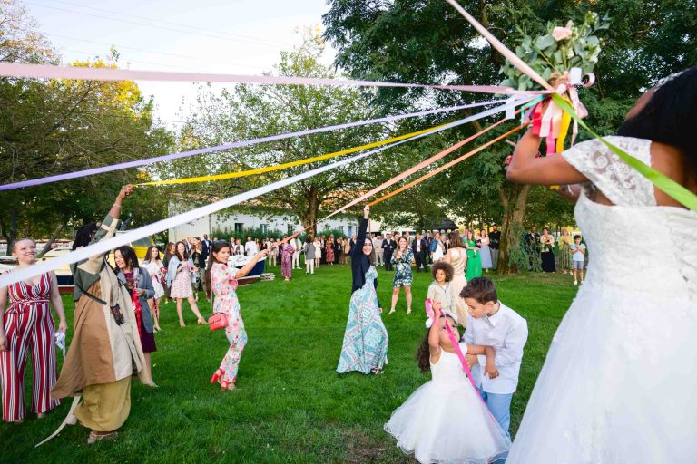 Lors d'un mariage Audenge, une mariée en robe blanche tient des rubans colorés avec un bouquet au bout. Les enfants et les invités, habillés de couleurs vives, tirent les rubans tout en acclamant une pelouse verte avec des arbres en arrière-plan. La scène festive est remplie de nombreux spectateurs sous un ciel clair et ensoleillé.