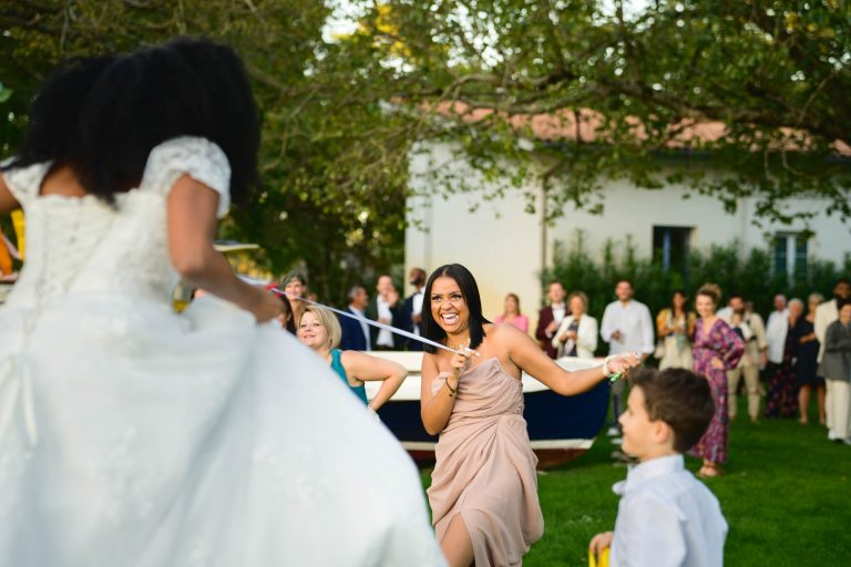 Une mariée en robe blanche saute à la corde sur une pelouse, tenant sa robe pour éviter de trébucher. Une femme en robe beige clair fait tourner la corde avec une expression joyeuse, tandis qu'un jeune garçon en chemise blanche et nœud papillon regarde. Une foule d'invités se tient à l'arrière-plan de ce magnifique Mariage Audenge, regardant et souriant.