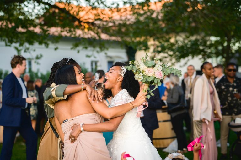 Une mariée en robe de mariée blanche et diadème, tenant un bouquet, serre joyeusement dans ses bras deux demoiselles d'honneur en robes beige clair lors de son Mariage Audenge. Elles sont à l'extérieur avec des arbres verts et un bâtiment blanc en arrière-plan. Des invités dans diverses tenues sont rassemblés autour, certains applaudissent et sourient, profitant de la célébration.