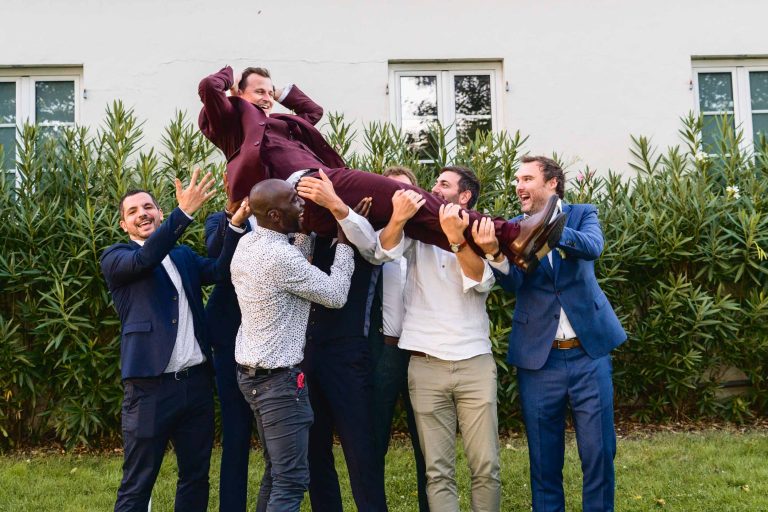 Un groupe de sept hommes, habillés pour la plupart en costume, soulèvent joyeusement un autre homme portant un costume marron et souriant largement. Ils se tiennent en plein air à Audenge, devant un bâtiment blanc avec des buissons verts et deux fenêtres. L'atmosphère festive suggère qu'il pourrait s'agir d'une célébration de mariage.