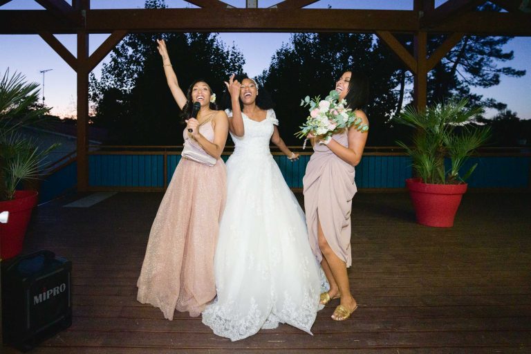 Trois femmes se tiennent joyeusement sur une terrasse en bois au coucher du soleil. La femme au centre, célébrant son mariage Audenge, porte une robe de mariée blanche et tient un bouquet. La femme à gauche, en robe rose, tient un micro, levant la main avec enthousiasme. La femme à droite, en robe beige, sourit.