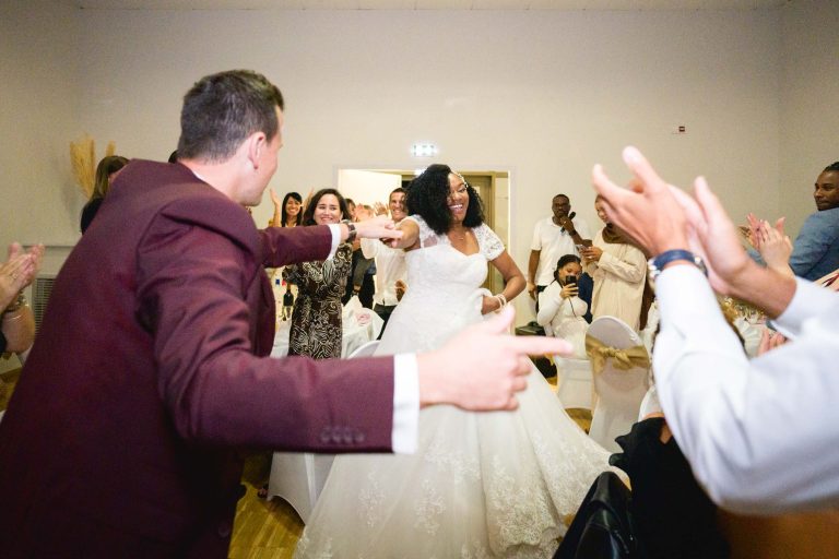 Une mariée en robe blanche danse joyeusement et sourit à son mariage Audenge, entourée d'invités qui l'applaudissent. Un homme en veste marron la désigne avec enthousiasme. D'autres invités, certains assis et d'autres debout, applaudissent et acclament en arrière-plan lors de la réception de mariage.