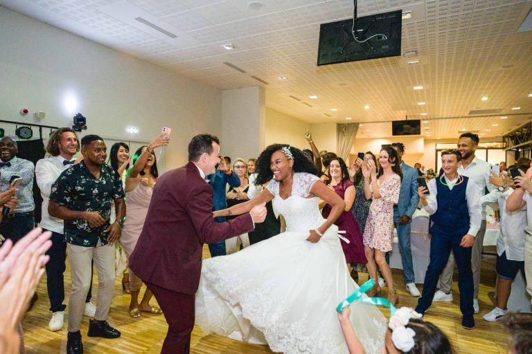 Une mariée et un marié dansent joyeusement lors de leur réception de mariage au Mariage Audenge, entourés d'invités enthousiastes, certains applaudissant tandis que d'autres prennent des photos et des vidéos. La mariée porte une robe blanche et le marié est en costume marron. La scène animée se déroule dans un lieu moderne et bien éclairé avec du parquet.