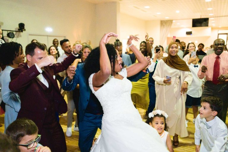 Un couple de mariés danse avec énergie lors de leur réception de mariage au Mariage Audenge, entourés d'invités enthousiastes qui applaudissent, acclament et filment la célébration avec leurs téléphones. La mariée porte une robe blanche et il y a des enfants au premier plan, dont l'un avec une couronne de fleurs et un autre en chemise blanche.