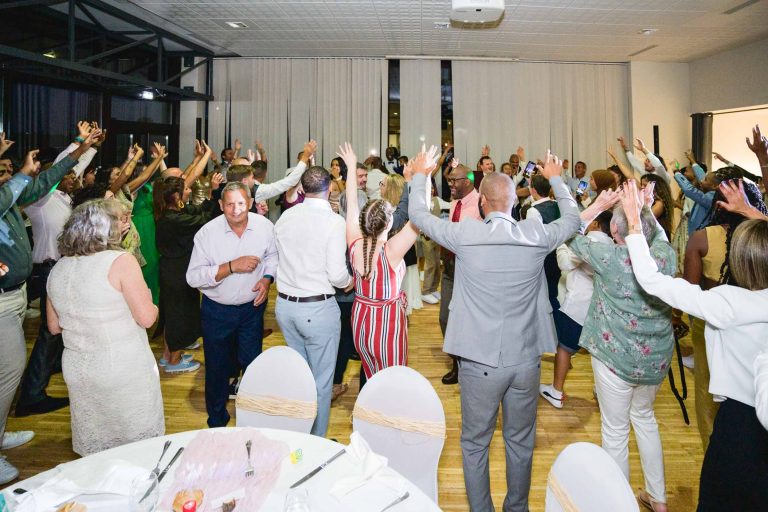 Un grand groupe de personnes est réuni dans une salle, toutes les mains levées et semblant profiter d'un événement festif du Mariage Audenge. Ils sont habillés de tenues semi-formelles ou décontractées, avec un mélange de vêtements à motifs et de couleurs unies. Il y a des tables à manger rondes avec des nappes blanches et des housses de chaises de couleur claire.