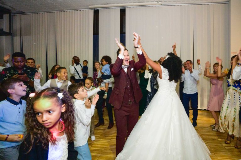 Une mariée en robe blanche et un marié en costume bordeaux dansent joyeusement au milieu d'une foule d'invités lors de leur mariage Audenge. Des personnes de tous âges les entourent, applaudissent et acclament. L'ambiance est animée et festive, les enfants se joignant à la fête. La salle est dotée de grandes fenêtres avec des stores blancs.