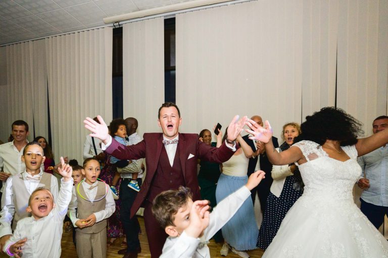 Une joyeuse célébration du Mariage Audenge à l'intérieur avec des gens qui dansent avec enthousiasme. Le marié, en costume bordeaux, et la mariée, en robe de mariée blanche, sont au centre, les bras levés. Les enfants et les invités autour d'eux sourient, applaudissent et acclament sous un éclairage doux et des stores verticaux blancs en arrière-plan.