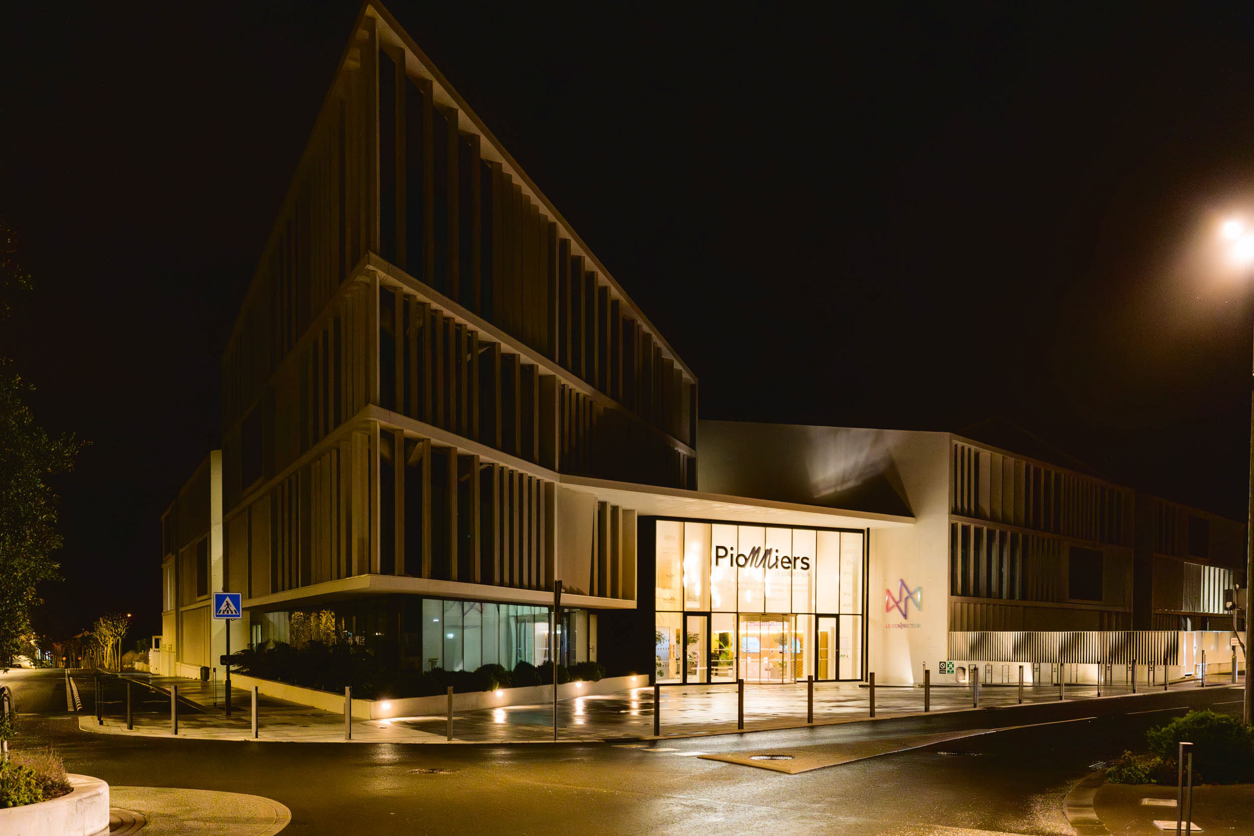 Un bâtiment moderne à l'architecture géométrique nette est illuminé la nuit. La façade est dotée de grandes baies vitrées et de surplombs angulaires. L'entrée, étiquetée « ProMilies », est bien éclairée. Un passage piéton est visible sur la gauche et des lampadaires illuminent les environs, une véritable source d'inspiration pour tout photographe d'architecture.