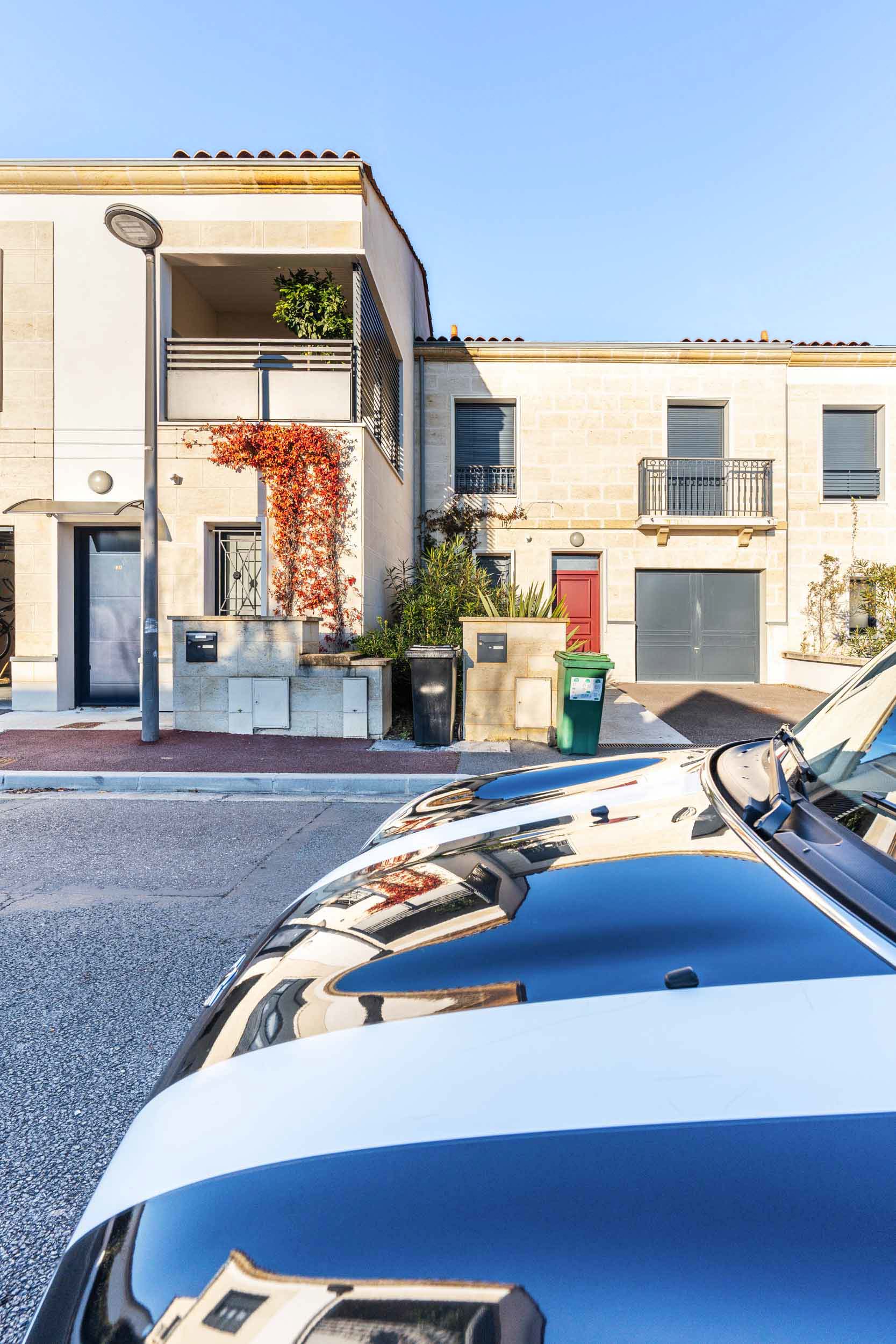 Une rue résidentielle avec des maisons beiges modernes à deux étages avec balcons et feuillage se dresse sous un ciel bleu. Au premier plan, une voiture noire avec des bandes blanches de course fait face aux maisons. À proximité, une poubelle verte et une poubelle noire de recyclage reposent sur le trottoir à côté d'un lampadaire, capturant l'œil de tout photographe d'architecture.