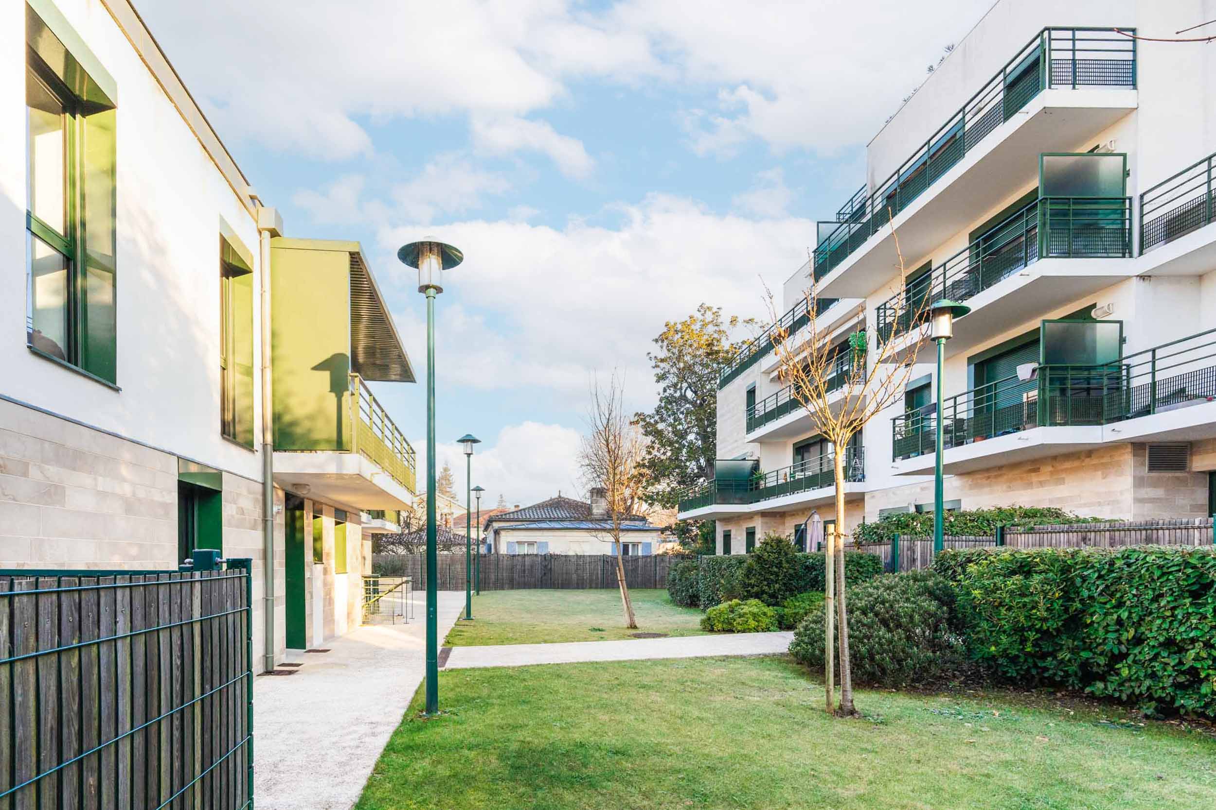 Un complexe résidentiel moderne composé de deux immeubles d'appartements à plusieurs étages avec balcons et façades végétalisées. Une cour gazonnée bien entretenue avec des arbres et des arbustes est située entre les bâtiments, parfaite pour un photographe d'architecture. Le ciel est partiellement nuageux et il y a des lampadaires le long d'une allée pavée.