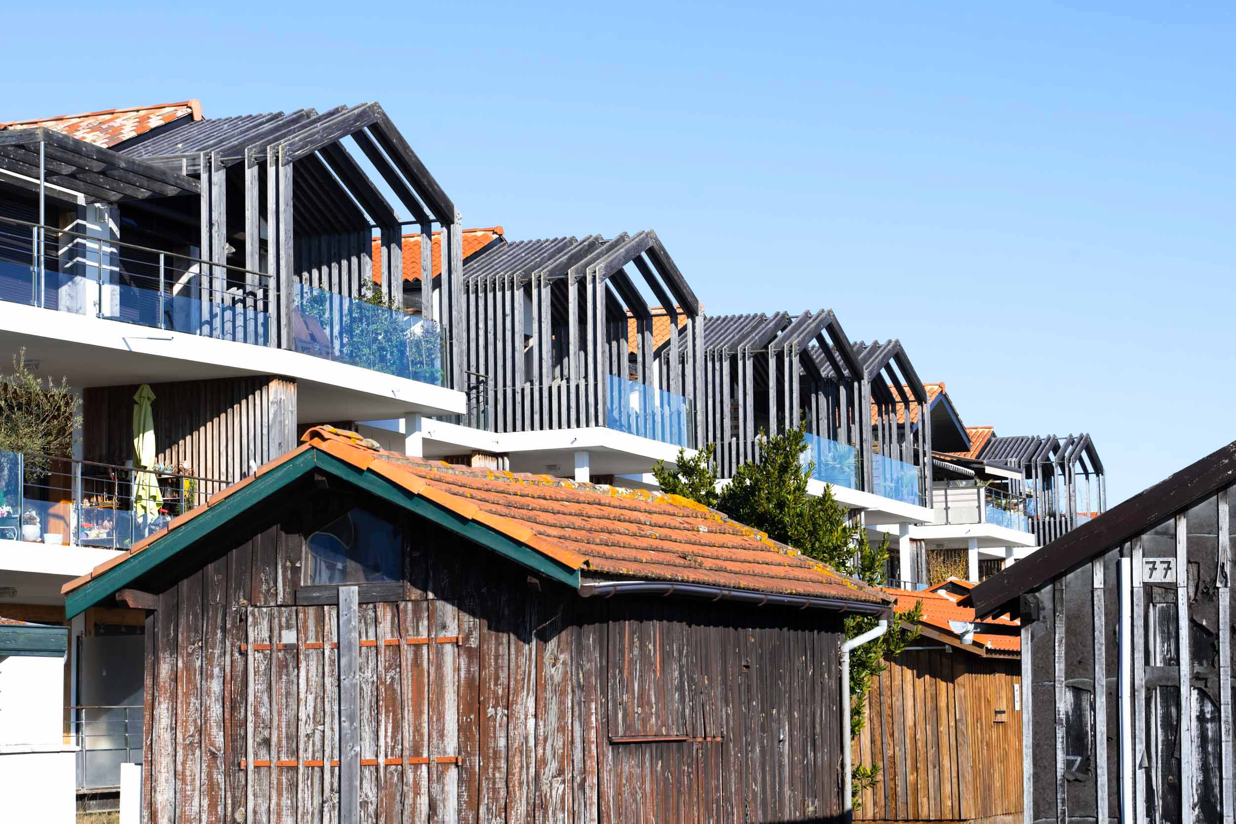 Derrière plusieurs hangars en bois rustiques aux toits de tuiles rouges se dresse une rangée d'appartements modernes aux façades en bois inclinées et aux grands balcons en verre. Le ciel bleu clair renforce le contraste architectural entre les nouvelles et les anciennes structures, tandis que les buissons et les arbres ajoutent un charme naturel. Idéal pour tout photographe d'architecture, ce décor allie à merveille design contemporain et caractère historique.