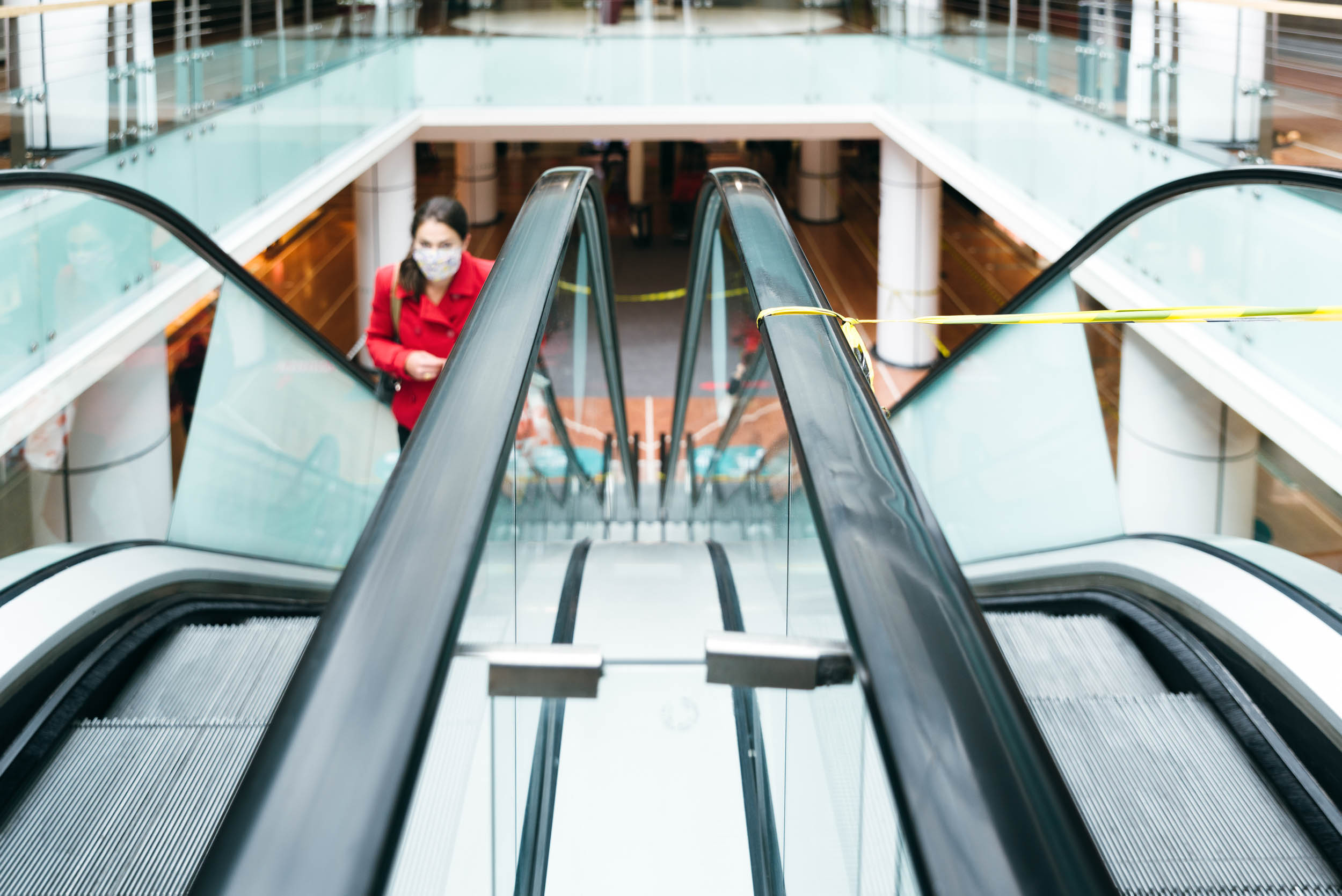 Une personne portant un manteau bordeaux et un masque facial monte un escalator dans un centre commercial couvert. Le centre commercial, capturé par un photographe d'architecture, est doté de balustrades en verre et de plusieurs étages. Un ruban d'avertissement jaune bloque l'escalator opposé. L'environnement semble moderne et bien éclairé.