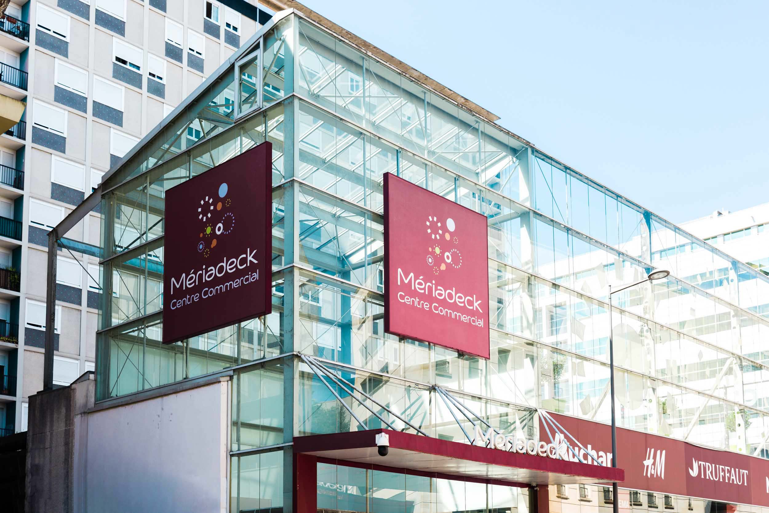 Un bâtiment moderne en verre avec deux grandes enseignes sur lesquelles est écrit « Mériadeck Centre Commercial ». Les enseignes sont marron avec du texte blanc et des motifs circulaires décoratifs. Parfaitement capturés par un photographe d'architecture, les panneaux élégants et l'entrée du bâtiment se démarquent, avec des structures plus hautes en arrière-plan bordeaux.