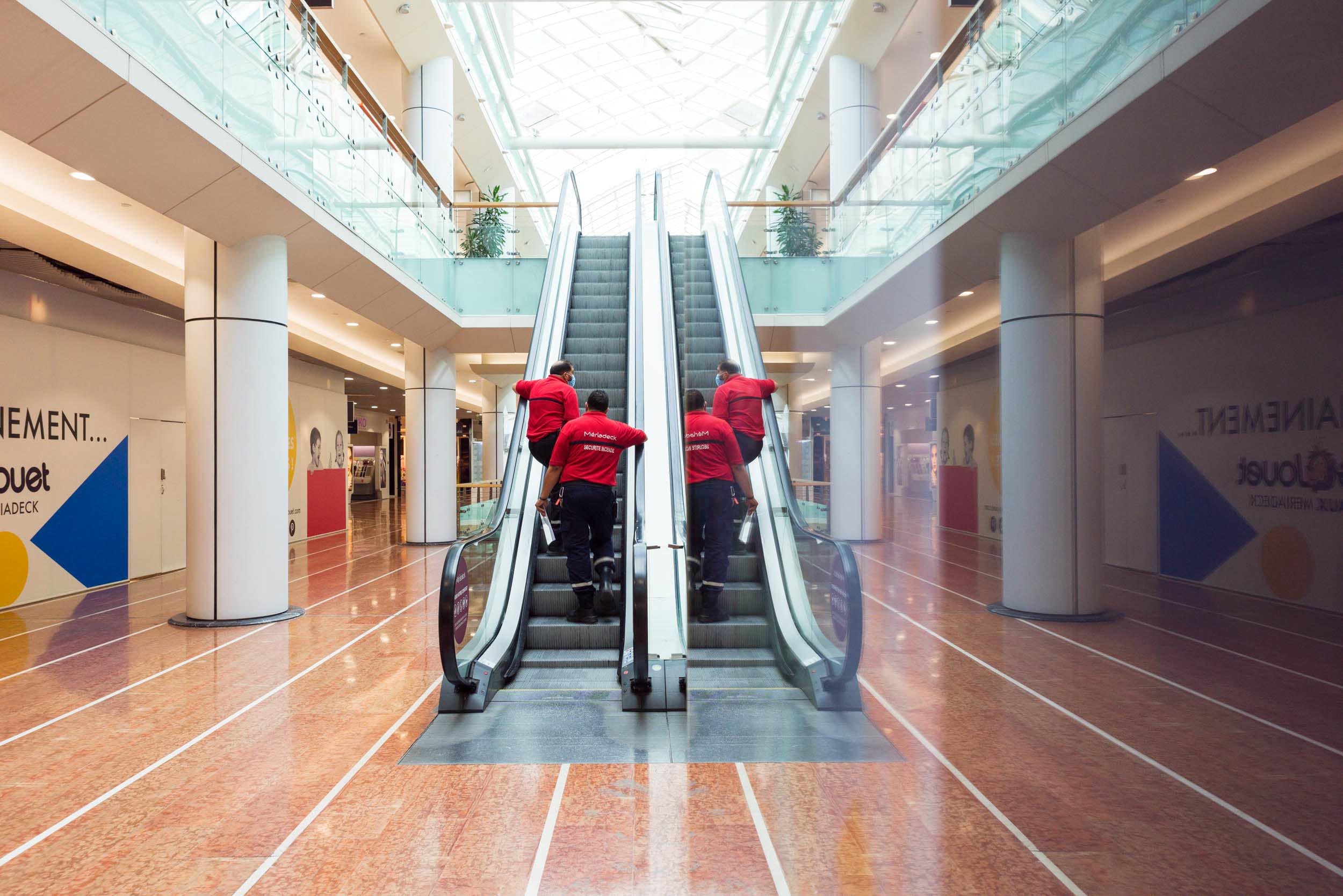 Trois personnes portant des vestes rouges montent un escalator dans un centre commercial spacieux et moderne, aux sols brillants et à l'éclairage intense. Le photographe d'architecture capture parfaitement la scène, mettant en valeur les grandes balustrades en verre et les hautes colonnes blanches. Des devantures de magasins aux volets roulants fermés et des publicités murales colorées bordent les deux côtés.