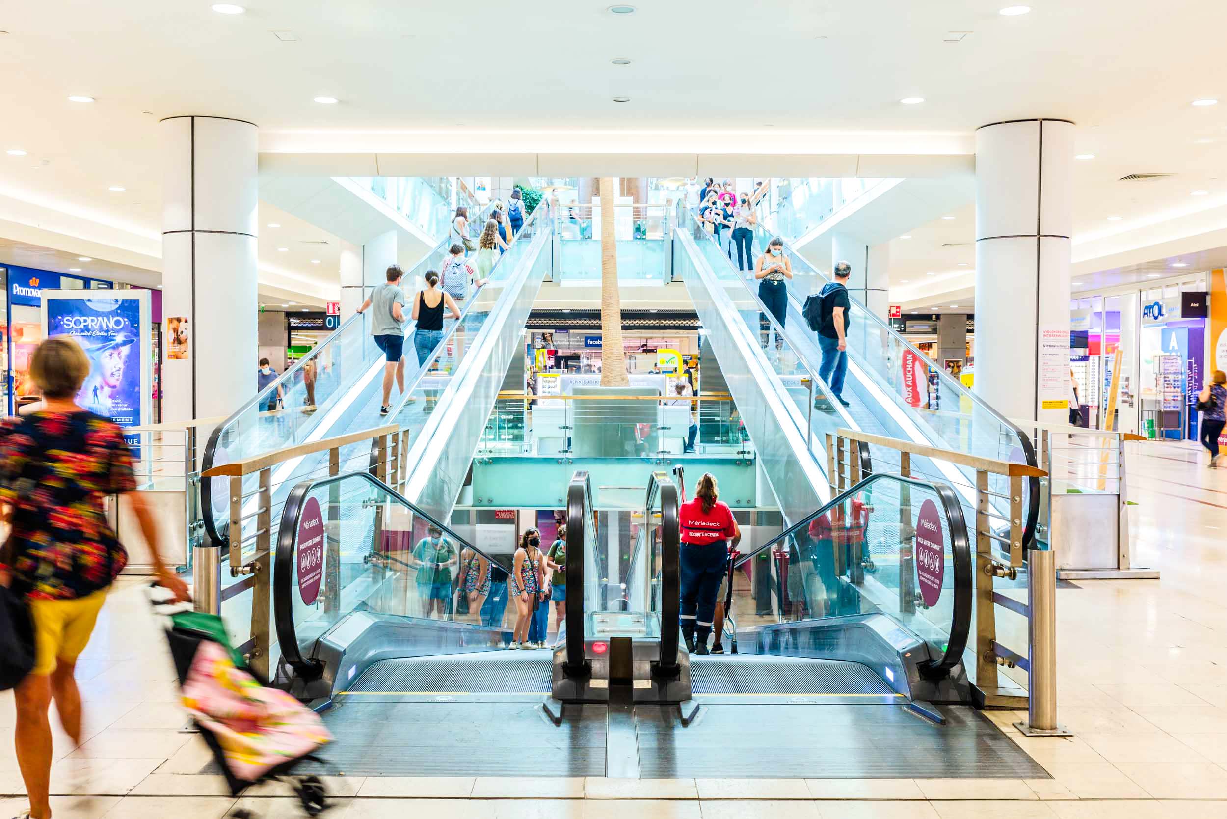 Un centre commercial très éclairé est doté de plusieurs escaliers mécaniques. On peut y voir des clients monter et descendre. Au premier plan, un individu pousse une poussette. Divers magasins aux enseignes et publicités colorées bordent l'arrière-plan, créant une atmosphère animée et dynamique que tout photographe d'architecture admirerait.