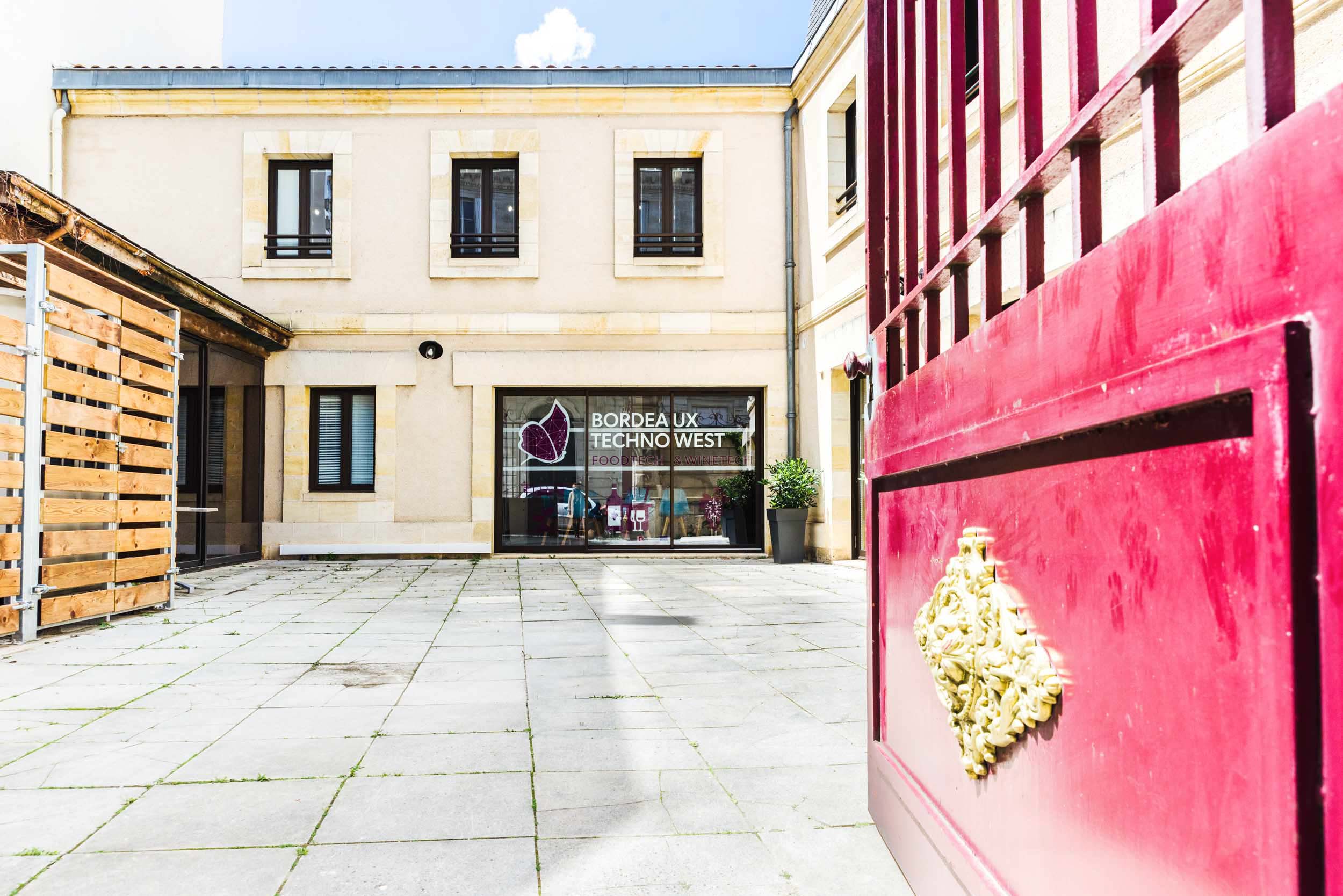 Une cour extérieure lumineuse au sol carrelé en pierre est visible à travers un portail rouge ouvert. Le bâtiment en arrière-plan, parfait pour un photographe d'architecture, porte un panneau indiquant « BORDEAUX TECHNO WEST ». Il présente des murs beiges, des fenêtres à cadre sombre et une structure en bois à gauche. Une plante en pot se trouve à droite.