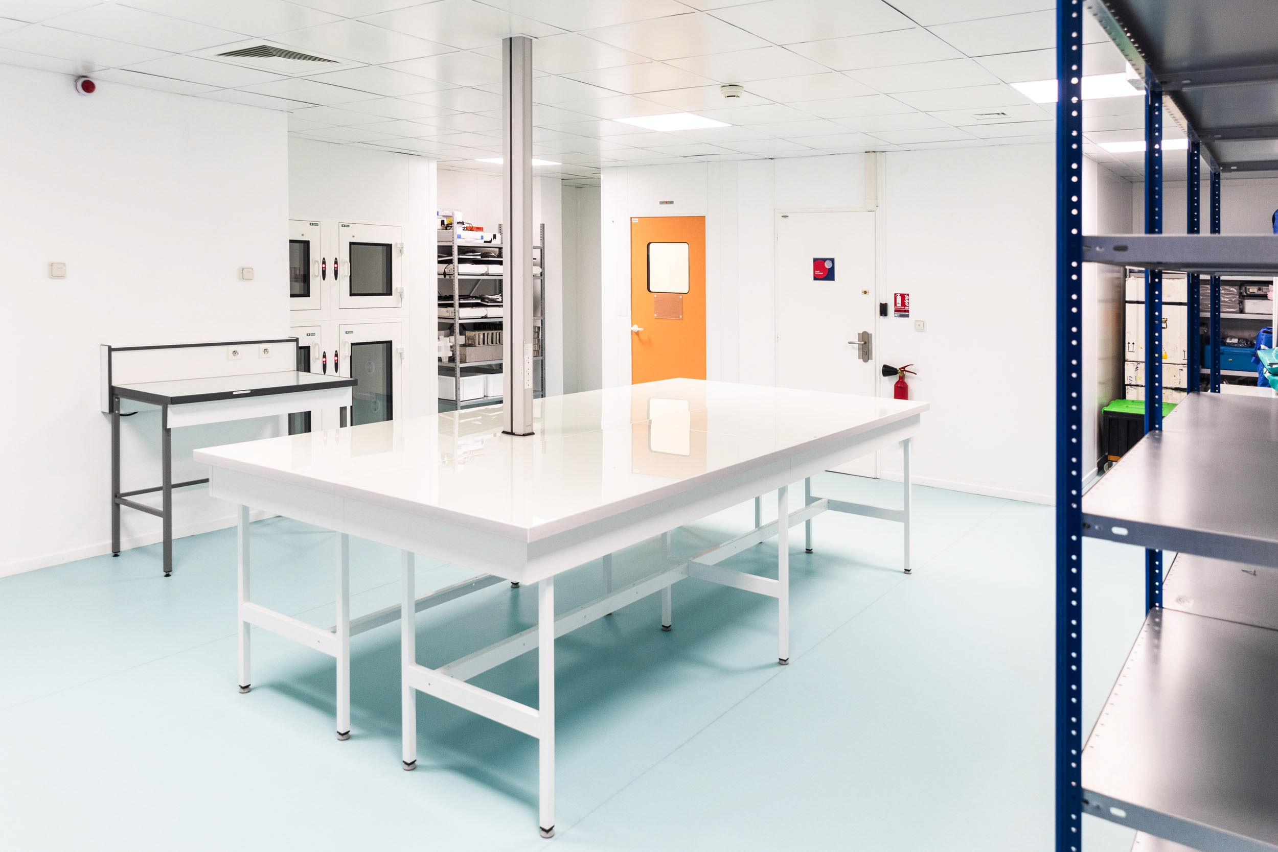 Une salle de laboratoire stérile aux murs et au plafond blancs. Une grande table propre au plateau blanc se dresse au centre, soutenue par des pieds en métal. Des étagères sont visibles à droite et une porte orange vif avec une petite fenêtre se trouve sur le mur du fond. Le sol est bleu clair et plusieurs postes de travail sont présents, créant une scène digne d'un photographe d'architecture.