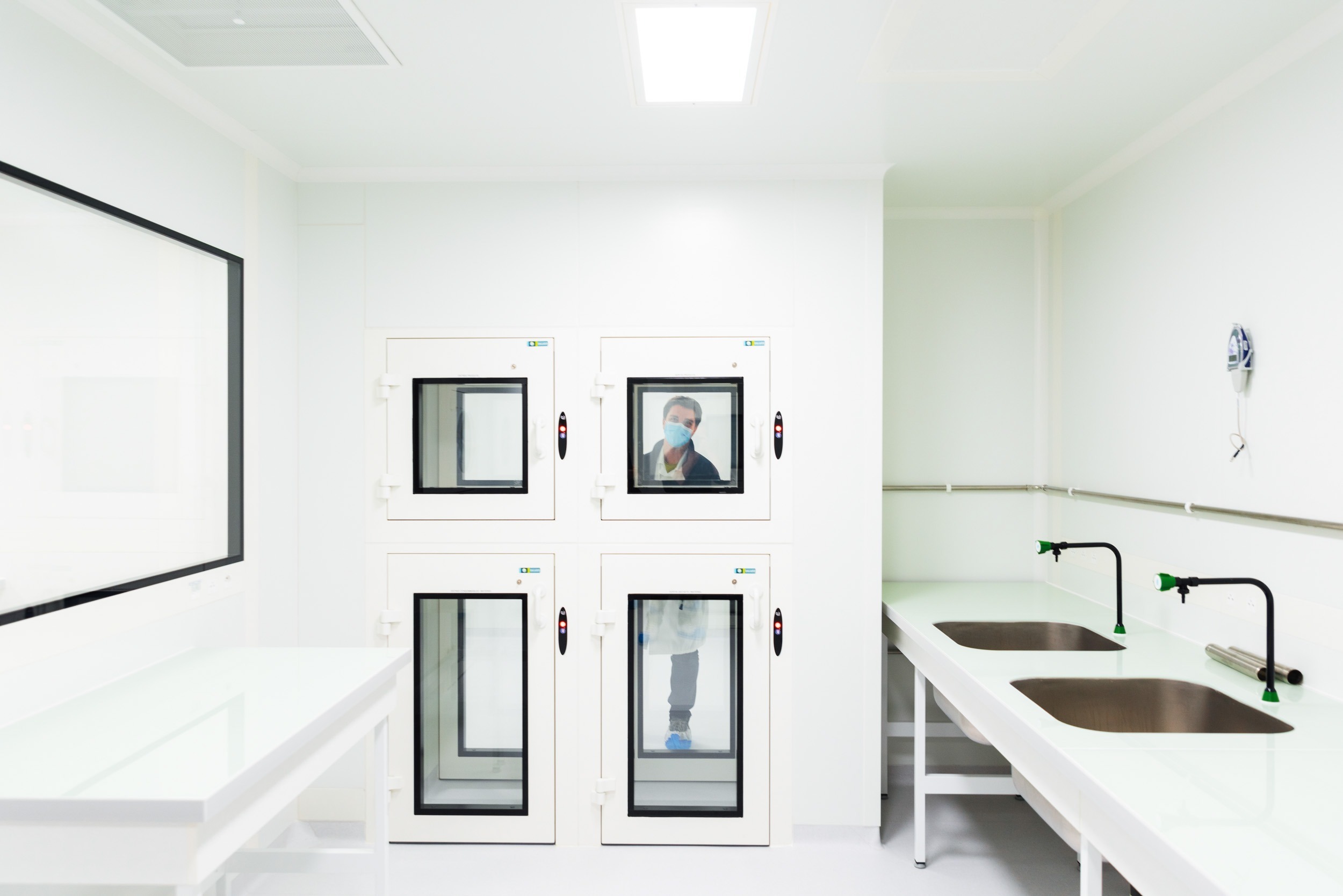 Une salle de laboratoire immaculée avec des murs et des comptoirs blancs, comme si elle avait été capturée par une photographie d'architecture. À gauche, quatre armoires à sas carrées et transparentes ont été installées, chacune avec une porte individuelle. Une personne en tenue de protection est visible à travers l'une des portes de l'armoire. À droite, il y a deux éviers en acier inoxydable.