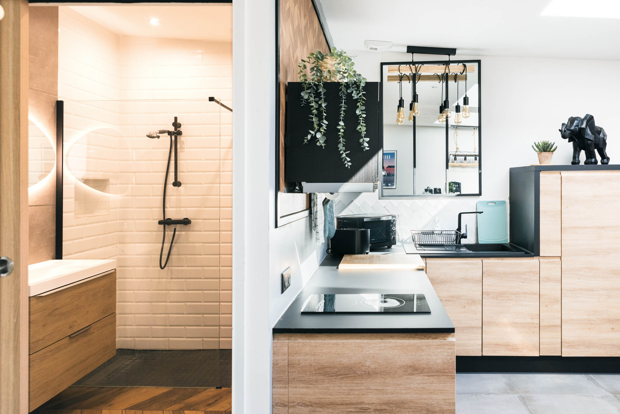 Une cuisine et une salle de bain modernes sont présentées, capturées par un photographe d'architecture. La salle de bain, à gauche, présente des carreaux de métro blancs, une vanité en bois et une douche noire. La cuisine à droite a des armoires en bois, des comptoirs noirs, une cuisinière, une plante suspendue et une figurine d'éléphant. Une fenêtre laisse entrer la lumière naturelle.