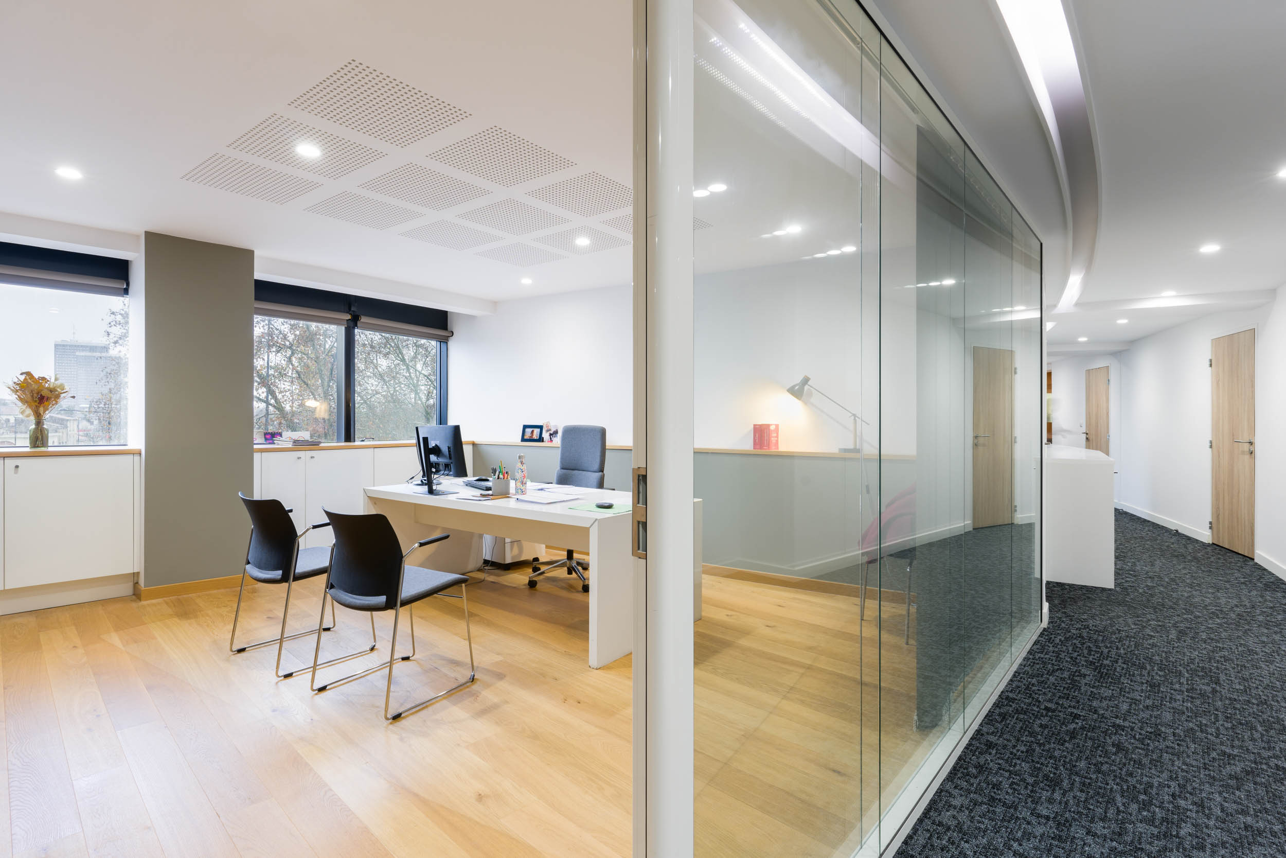 Espace de bureau moderne avec de grandes baies vitrées et une paroi vitrée séparant les pièces, conçu par un photographe d'architecture renommé. À l'intérieur, un bureau avec un ordinateur, deux chaises noires, une chaise de bureau grise et des plantes en pot. Un plancher en bois contraste avec le couloir sombre recouvert de moquette à l'extérieur. Un éclairage encastré offre une ambiance lumineuse et épurée.