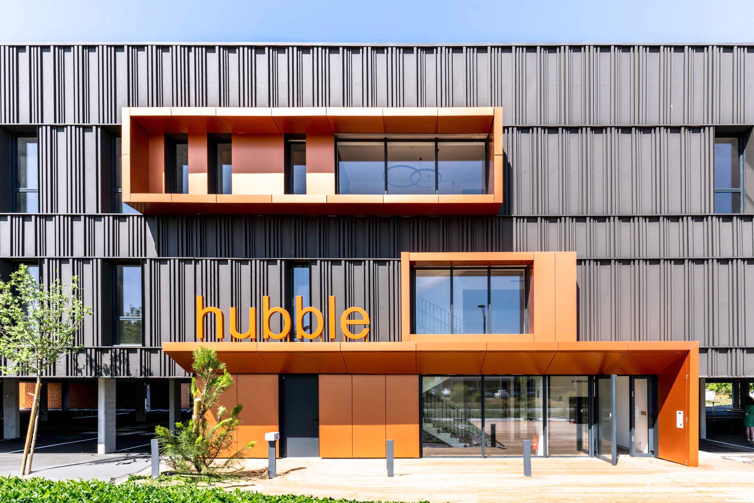 Un bâtiment moderne, surnommé « Hubble » sur sa façade, trône fièrement au sein du bâtiment inaugural de Mérignac. Il présente un extérieur sombre à motifs verticaux avec des touches d'orange. L'entrée comprend un cadre orange audacieux et une porte vitrée, entourée de petits arbres et de buissons sous un ciel bleu clair.