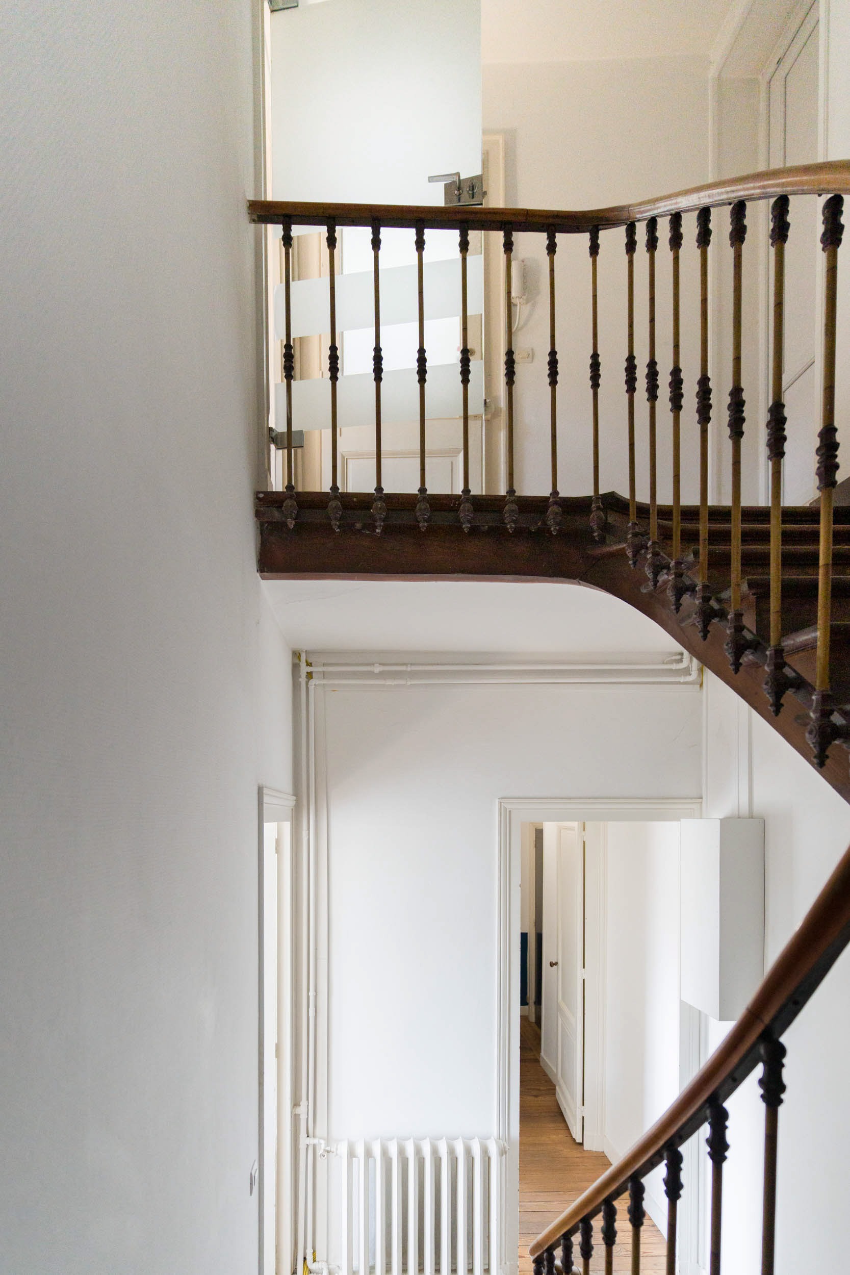 Un escalier étroit et bien éclairé, composé d'un escalier en bois avec rampes sombres et balustres en fuseaux ornés, s'incurve vers le haut à gauche, menant à un couloir supérieur. Ce décor à thème bordeaux est mis en valeur par des murs blancs et un radiateur en dessous, avec des portes ouvertes menant aux pièces du niveau inférieur.