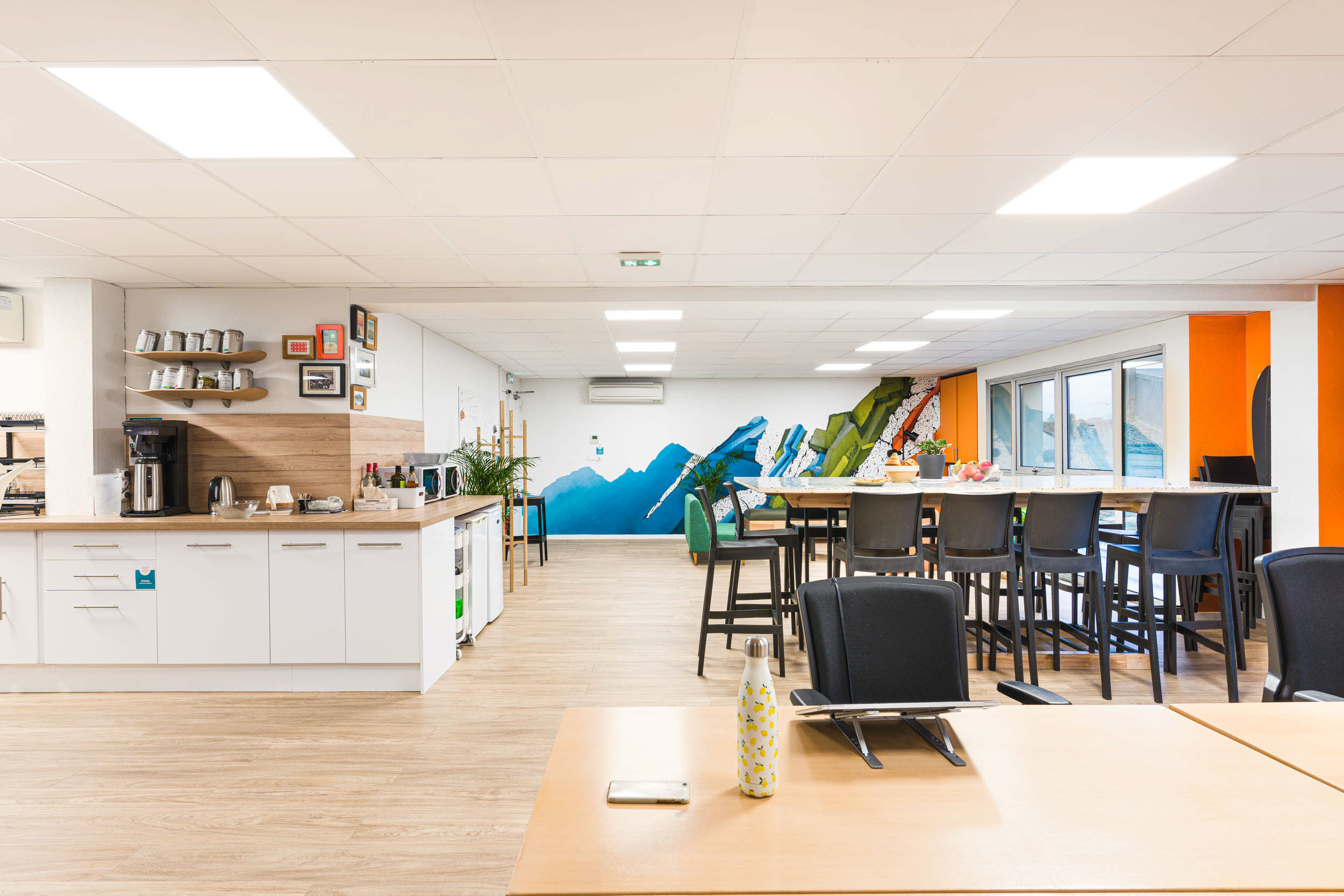 Une salle de pause lumineuse avec parquet et plafonds blancs. L'espace, soigneusement conçu dans le style de photographe d'architecture décoration d'intérieur bordeaux, comprend une kitchenette avec des armoires blanches, une cafetière et des étagères avec des tasses. Une longue table avec des chaises hautes fait face à une peinture murale colorée représentant des montagnes. De grandes fenêtres illuminent la pièce.