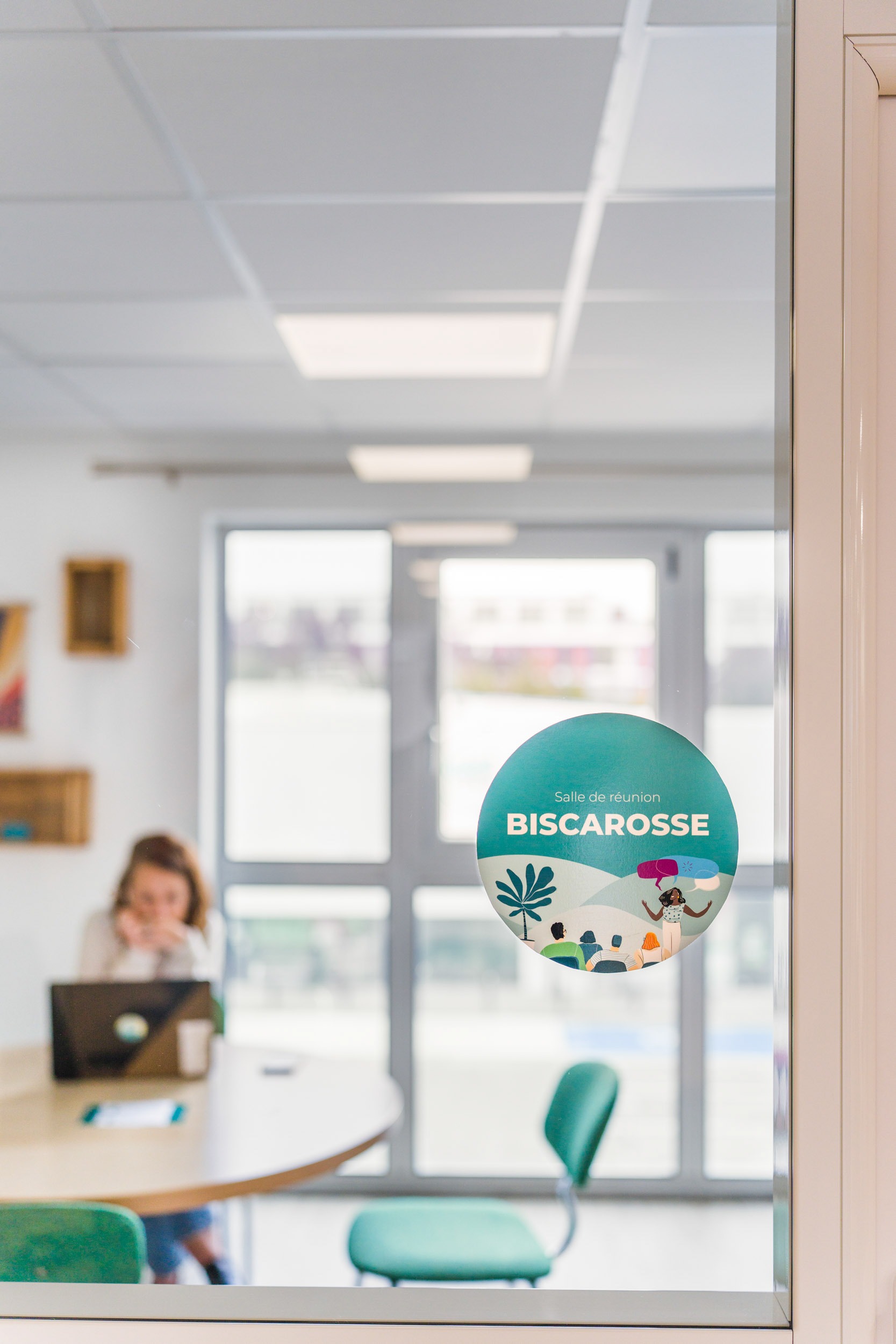 Une porte vitrée sur laquelle est écrit « Salle de réunion Biscarrosse » révèle une salle de réunion bien éclairée. À l'intérieur, une femme travaille sur un ordinateur portable à une table ronde entourée de chaises vertes. Des étagères décorées sont montées sur les murs blancs et de grandes fenêtres à l'arrière-plan laissent entrer une abondante lumière naturelle, incarnant une décoration d'intérieur exquise.