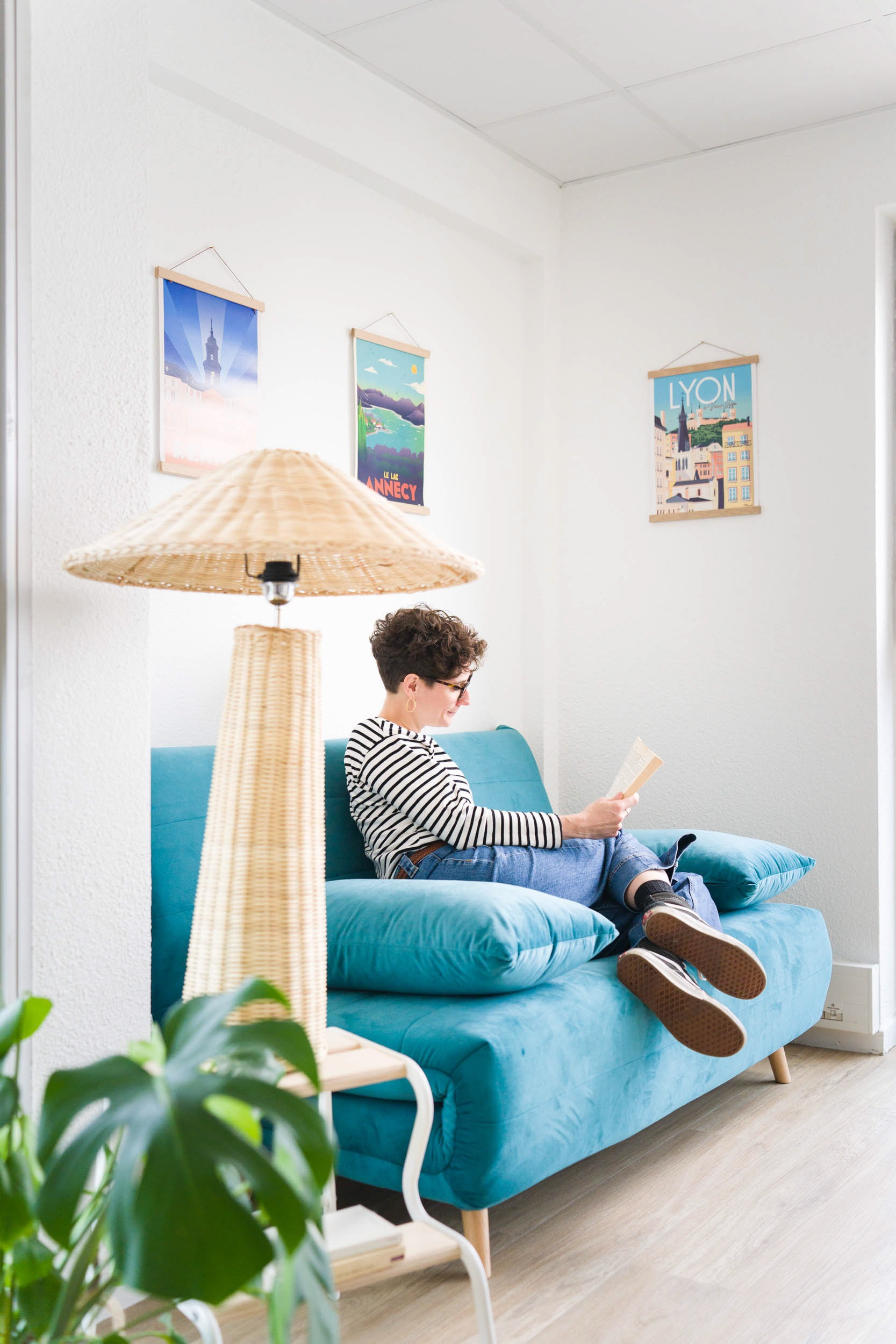 Une personne aux cheveux courts, portant des lunettes, une chemise rayée et un jean, est assise sur un canapé turquoise en train de lire un livre. Un grand lampadaire avec un abat-jour en osier se trouve à proximité. Le mur derrière le canapé est orné de trois affiches encadrées. Une plante verte est visible au premier plan, ce qui ajoute à l'ambiance de photographe d'architecture.