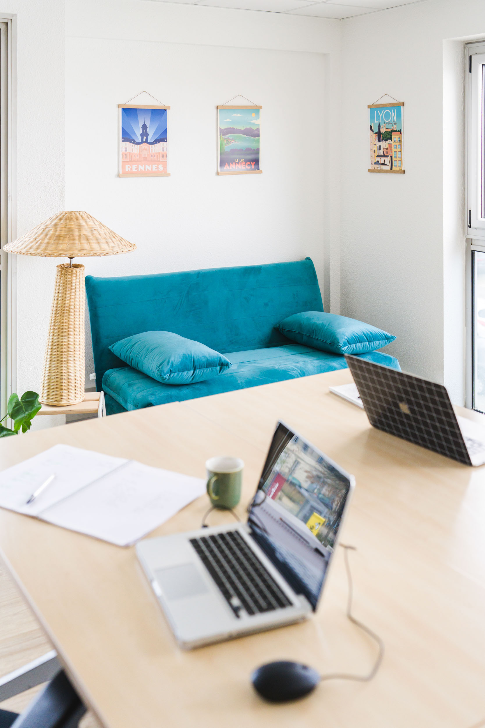 Un espace de bureau lumineux au cœur de Bordeaux comprend une table en bois avec deux ordinateurs portables, un carnet ouvert, un stylo et une tasse à café. Une lampe en osier et un canapé bleu canard avec des coussins assortis créent un coin confortable. Trois affiches colorées de la ville sont suspendues au-dessus du canapé, complétées par une plante verte pour une touche de décoration d'intérieur parfaite.