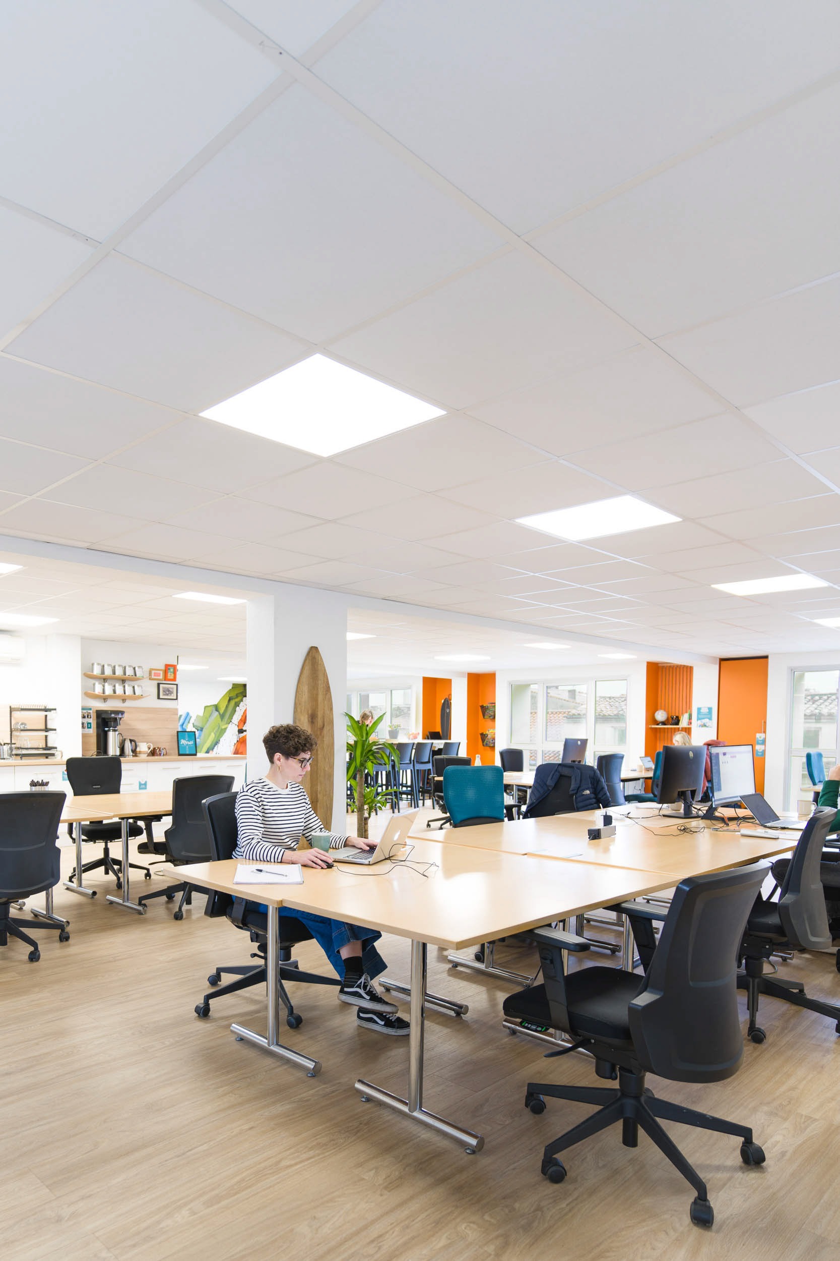 Un bureau spacieux, lumineux et ouvert avec des meubles modernes conçus par un photographe d'architecture renommé. Une personne est assise seule à un grand bureau travaillant sur un ordinateur portable. La pièce comprend plusieurs bureaux avec des chaises ergonomiques, une planche de surf placée de manière décorative en arrière-plan et des murs d'accent orange ajoutant une touche de couleur.