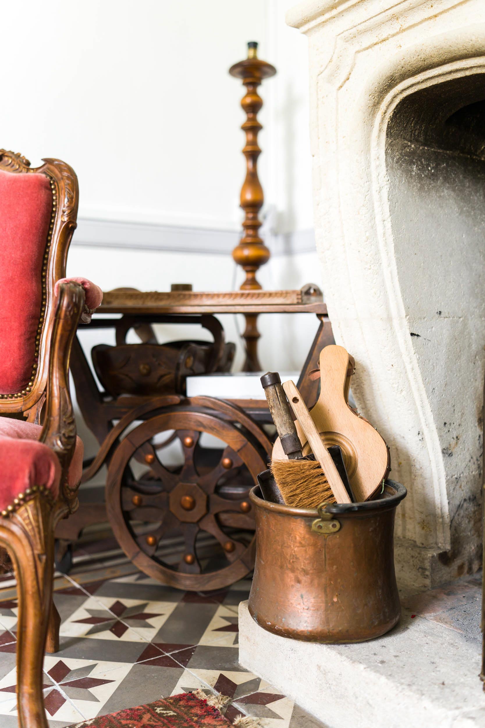 Un salon vintage comprend un fauteuil bordeaux rembourré, une table d'appoint en bois avec des roulettes décoratives et une cheminée en pierre à l'ancienne. À côté de la cheminée se trouve un seau en cuivre contenant un petit balai, un instrument de musique en bois et d'autres objets divers. Le sol présente un motif de carrelage, parfait pour toute décoration d'intérieur.