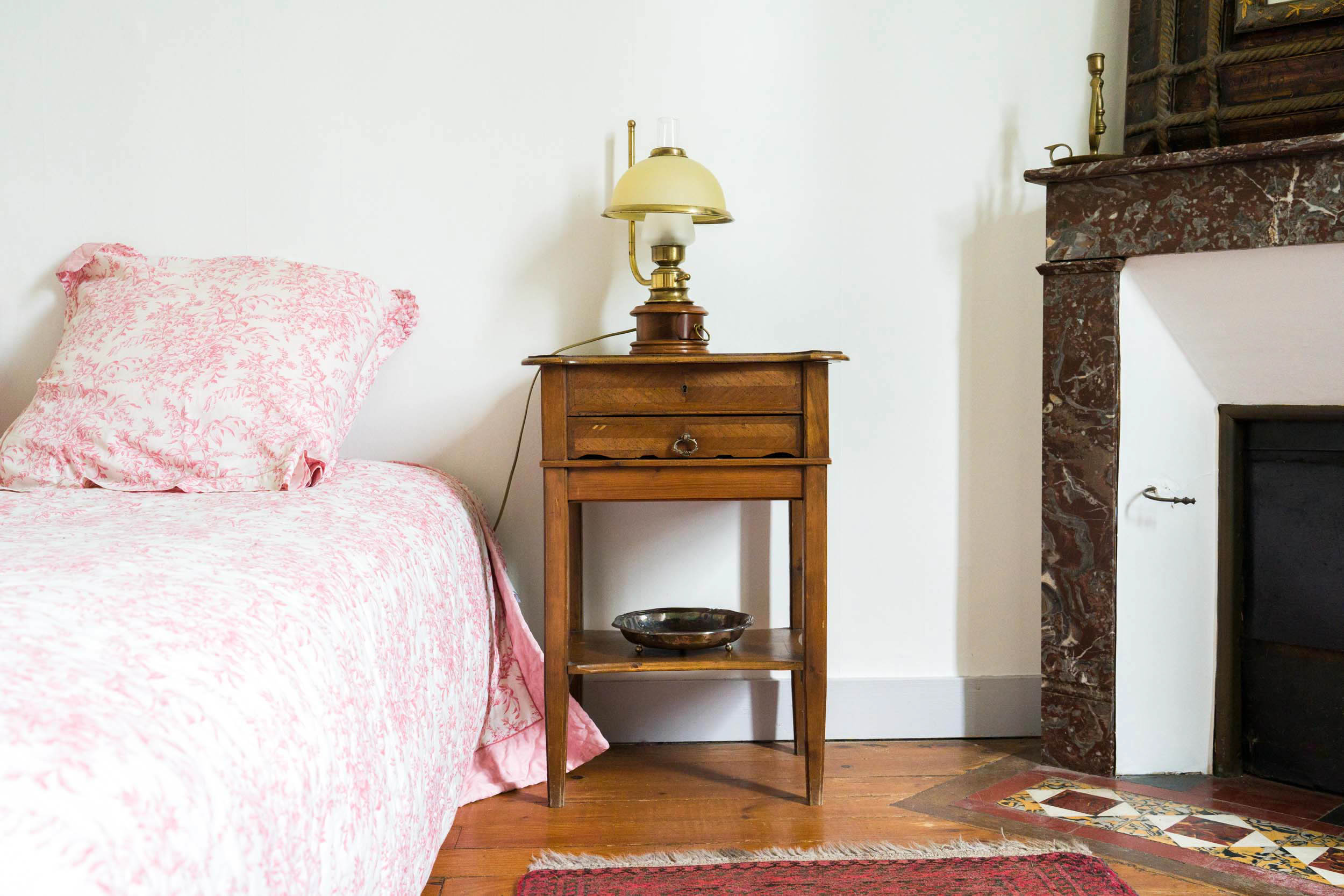 Une chambre confortable est dotée d'une table de nuit en bois avec une lampe et un bol vintage. La table de nuit se trouve entre un lit avec une literie à fleurs roses et une cheminée en marbre bordeaux. Capturée par l'œil d'un photographe d'architecture, la pièce respire la chaleur avec son parquet en bois et son tapis à motifs au premier plan.