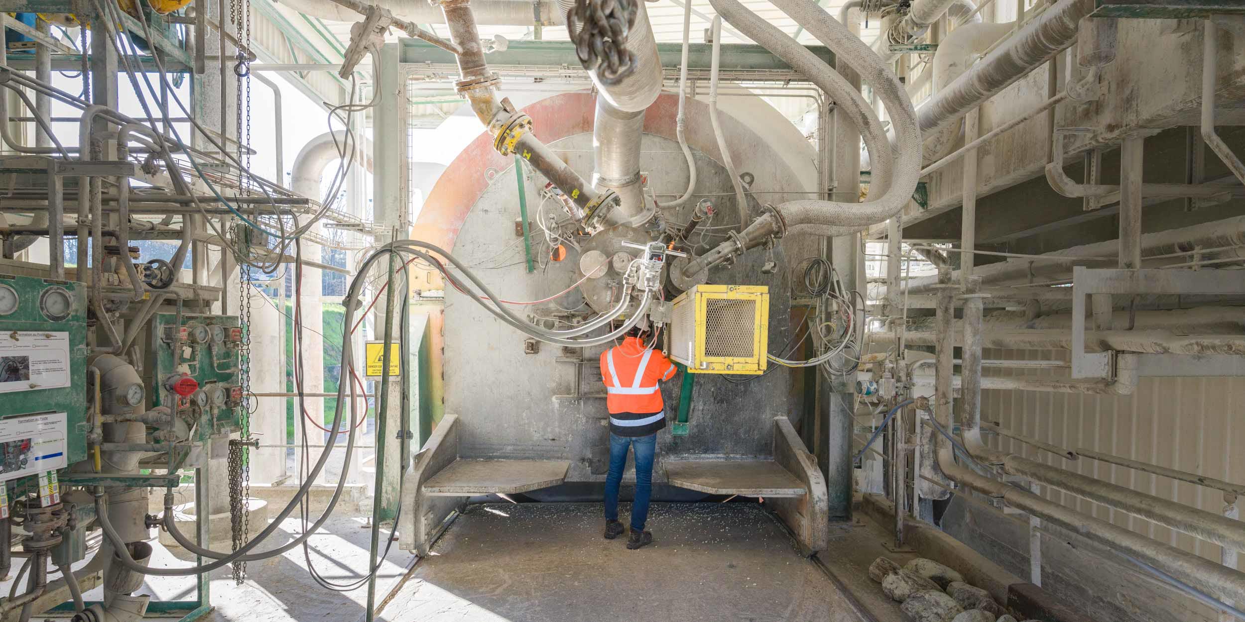 Un ouvrier portant un gilet de sécurité orange vif fait fonctionner une grande machine industrielle dans un décor d'usine, rappelant la précision méticuleuse que l'on retrouve dans la photographie d'architecture. La machine est entourée de nombreux câbles, tuyaux et structures métalliques. La lumière du jour filtre par une fenêtre au-dessus, illuminant la scène.