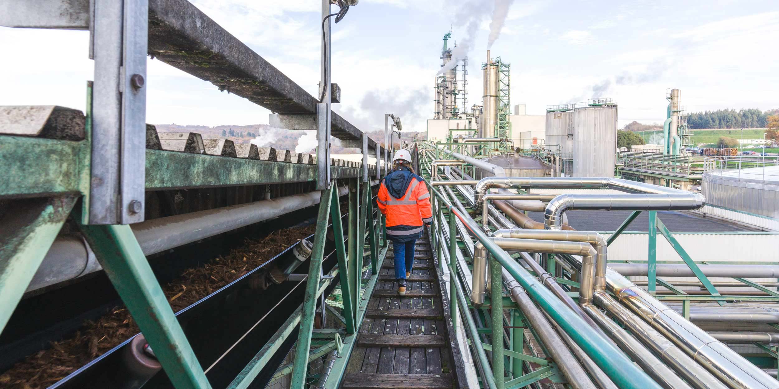 Un ouvrier portant un casque de sécurité blanc et un gilet de sécurité orange vif marche le long d'une étroite passerelle métallique, entourée de tuyaux et de machines industrielles dans une usine de fabrication. De la fumée s'élève des hautes cheminées en arrière-plan, avec un paysage d'arbres et de ciel visible à l'horizon ressemblant à une photographie d'architecture industrielle.