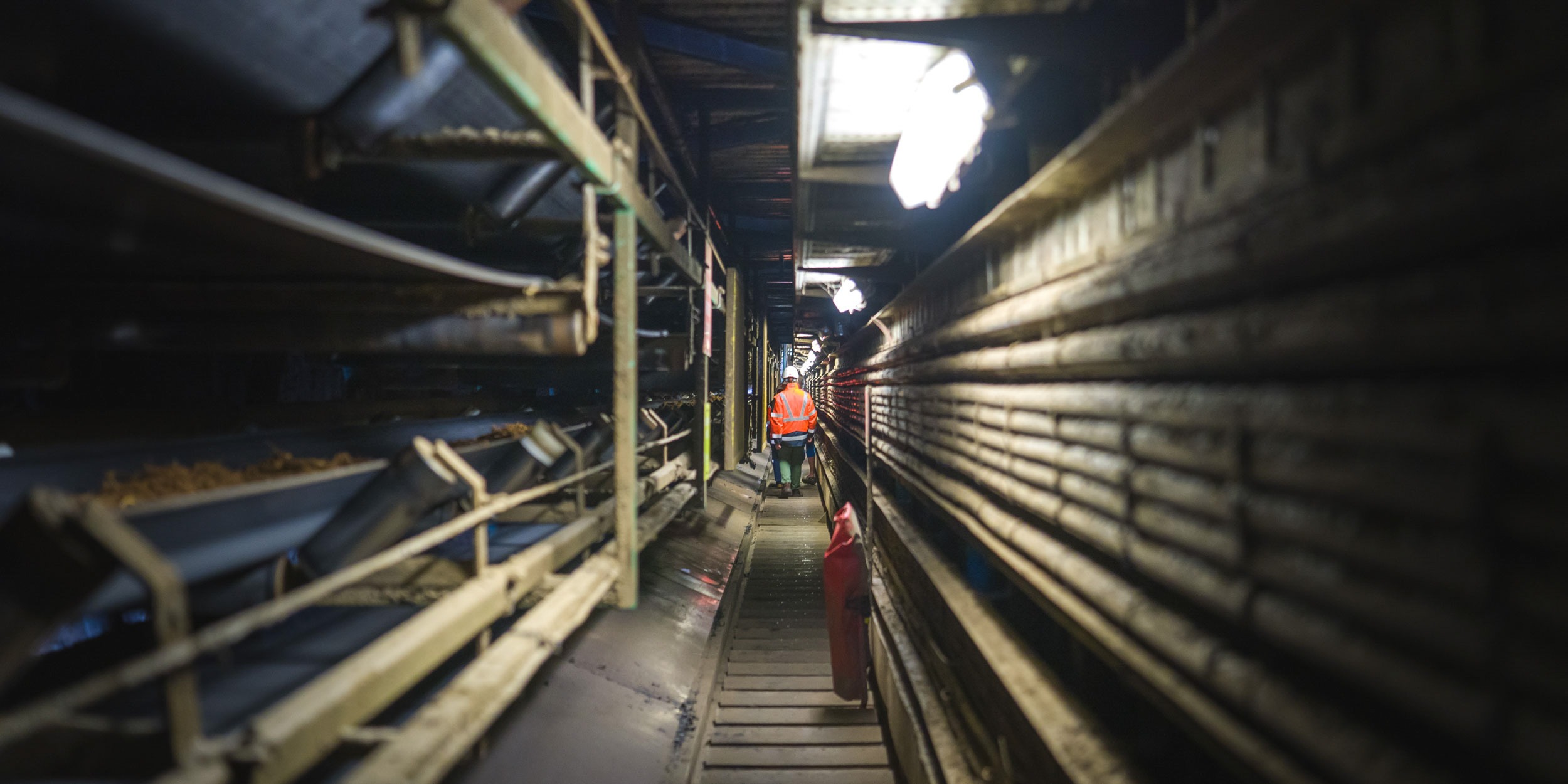 Un homme vêtu d'une veste bordeaux marche dans une allée industrielle étroite et faiblement éclairée, bordée de machines et de tuyaux. L'individu se trouve au milieu de l'allée, entouré de tapis roulants transportant des matériaux, sous une lumière zénithale qui projette une lueur crue.