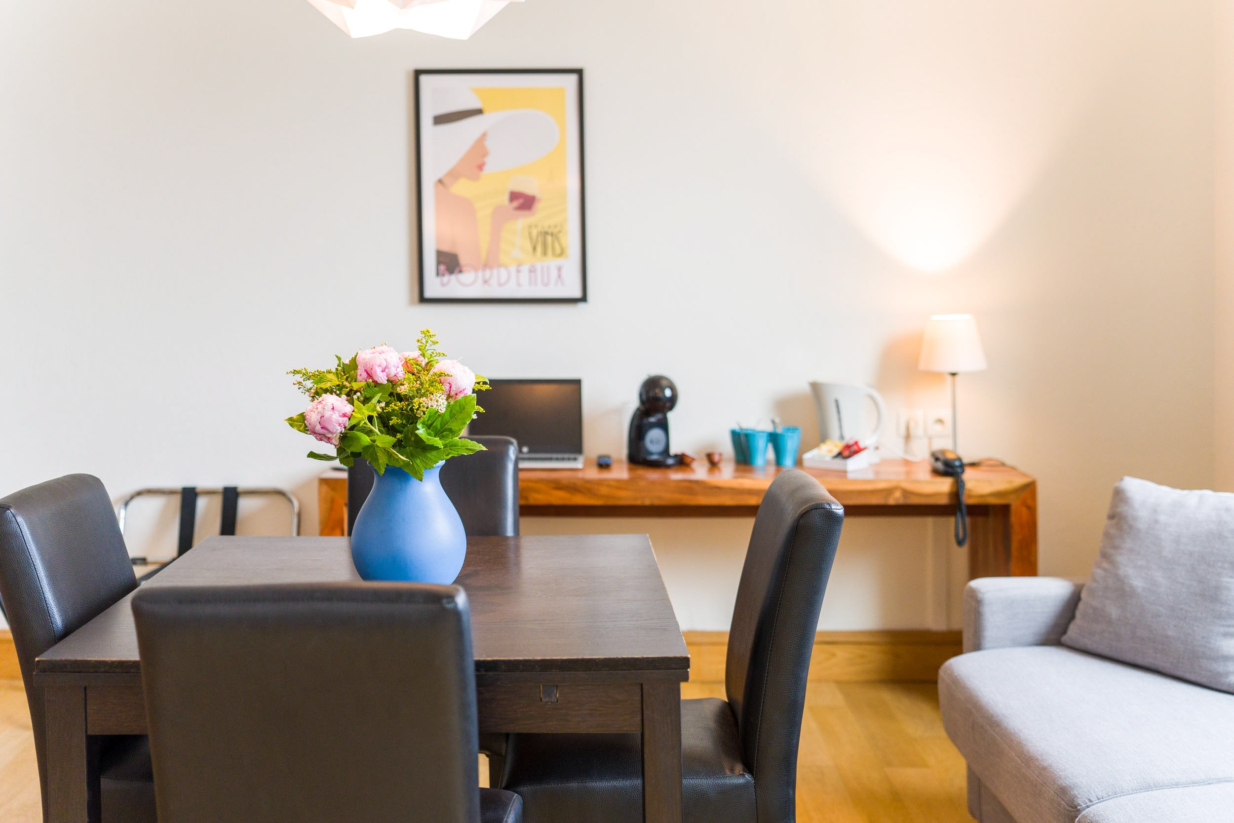 Une pièce chaleureuse conçue par un talentueux photographe d'architecture comprend une table à manger en bois avec quatre chaises noires. Un vase bleu avec des fleurs roses est posé sur la table. En arrière-plan, un bureau en bois avec une machine à café noire, des tasses bleues, une bouilloire et une lampe. Une affiche vintage fixée au mur et un canapé gris sont également visibles.