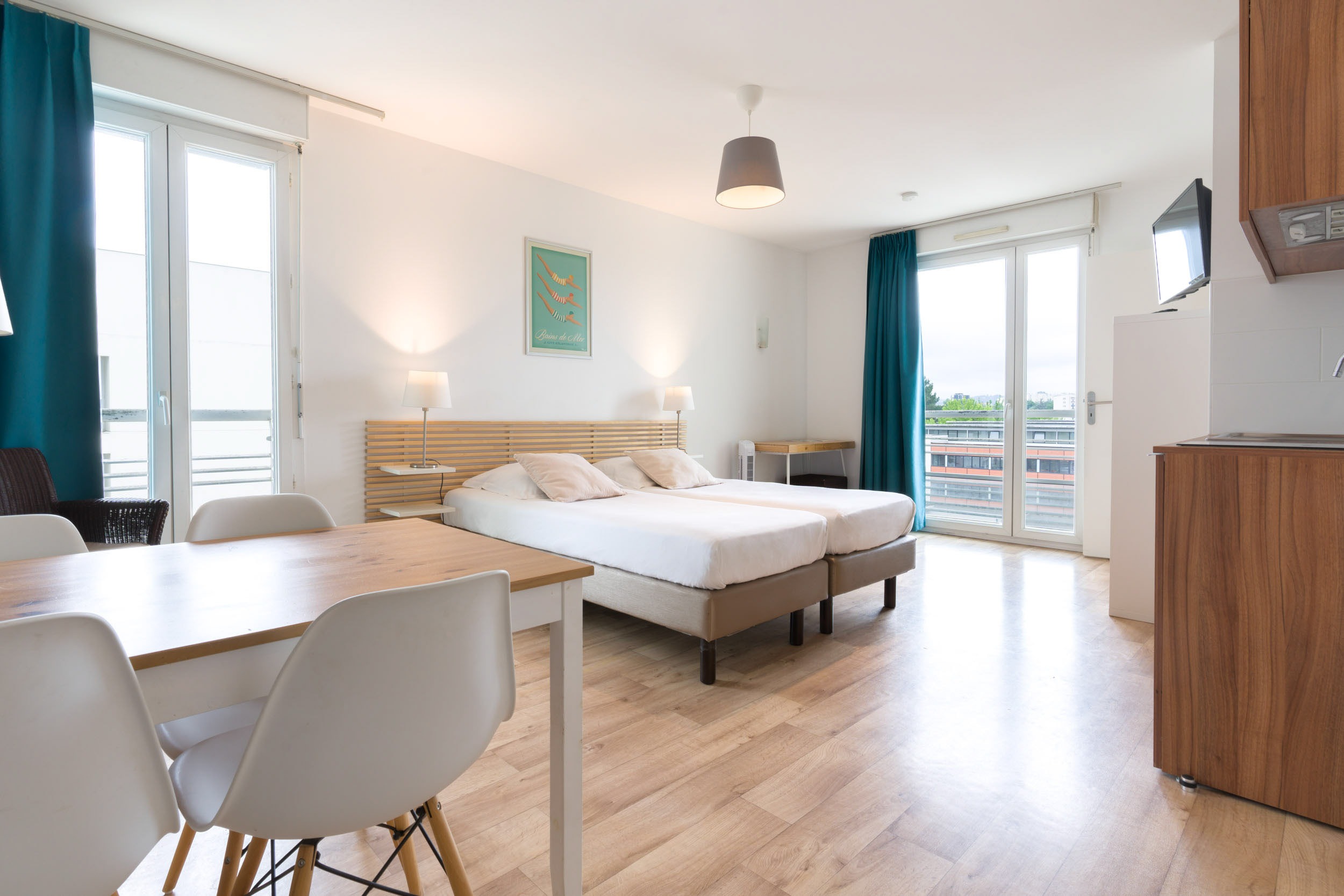 Cette chambre d'hôtel moderne et lumineuse avec parquet dispose d'un lit double avec une tête de lit en bois, d'une télévision murale et d'une petite table à manger avec des chaises blanches. La décoration intérieure de la chambre capture l'essence des teintes bordeaux sophistiquées. De grandes fenêtres avec des rideaux bleus offrent une lumière naturelle et une vue sur les bâtiments voisins.