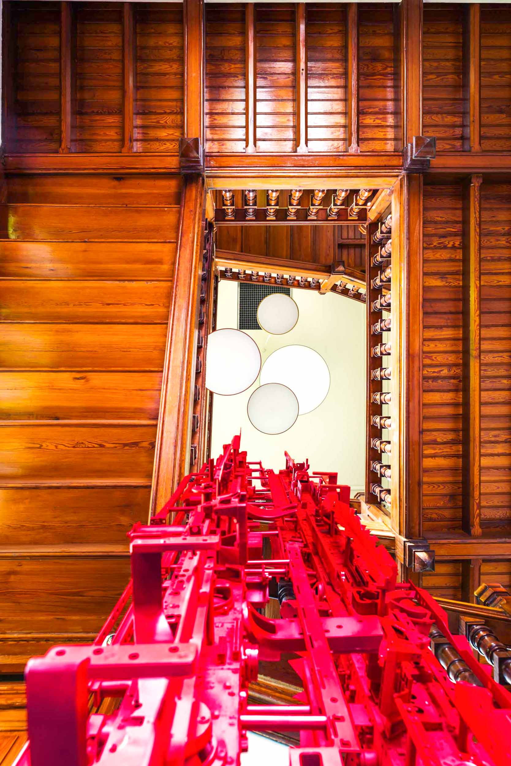 Vue plongeante sur plusieurs niveaux d'un bâtiment aux poutres et balustrades en bois, mettant en valeur une structure bordeaux complexe qui s'étend verticalement le long du centre. Des luminaires blancs circulaires sont visibles au sommet, contrastant avec les tons chauds du bois et l'installation rouge frappante - parfait pour tout photographe d'architecture passionné de décoration d'intérieur.