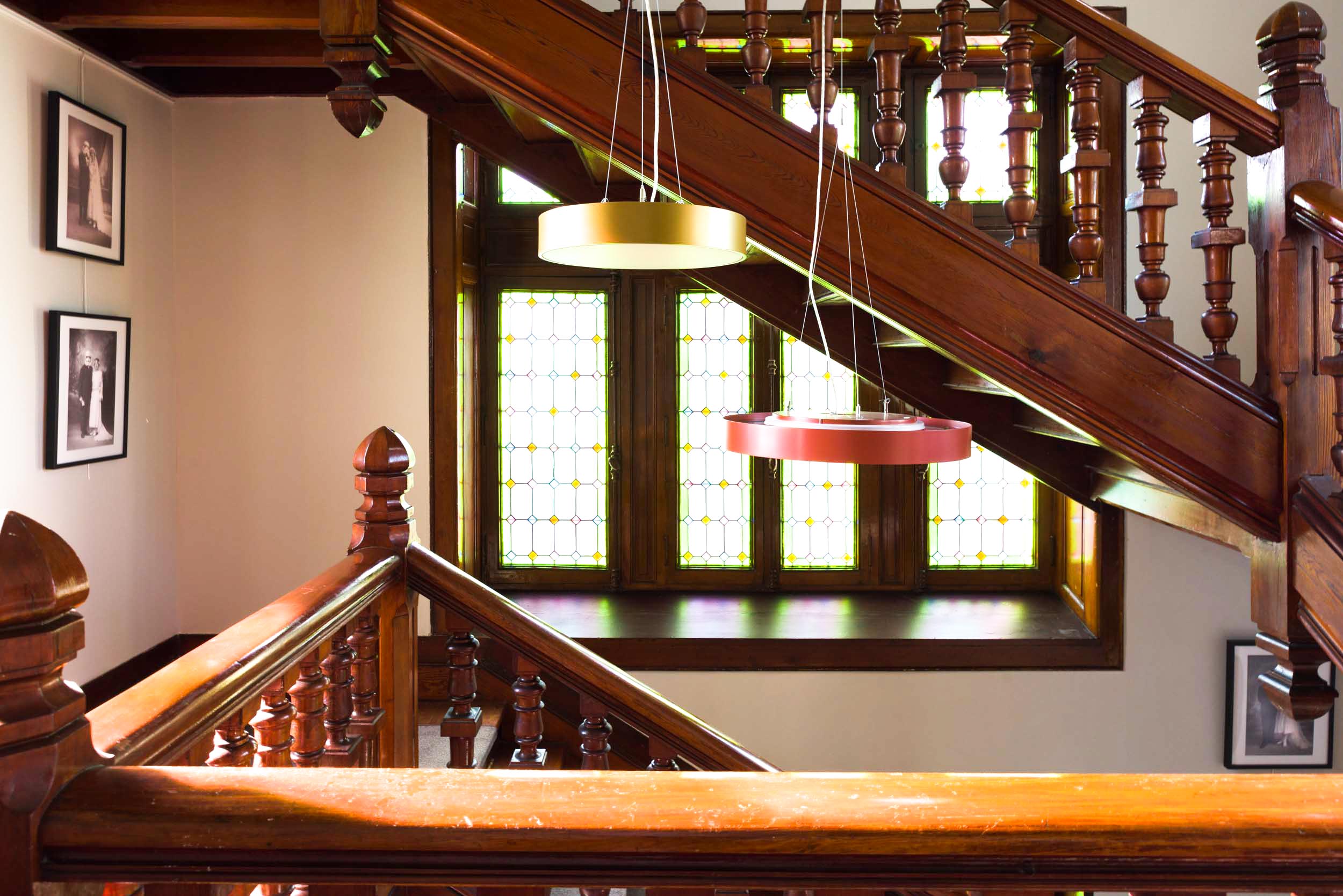 Un escalier baigné de lumière, conçu par un célèbre photographe d'architecture et de décoration d'intérieur de Bordeaux, présente des balustres et des rampes en bois complexes. Trois suspensions circulaires sont suspendues au-dessus, une dorée et deux rouges. Des vitraux à motifs verts et jaunes au niveau supérieur laissent filtrer une douce lumière. Des photographies en noir et blanc encadrées ornent les murs.