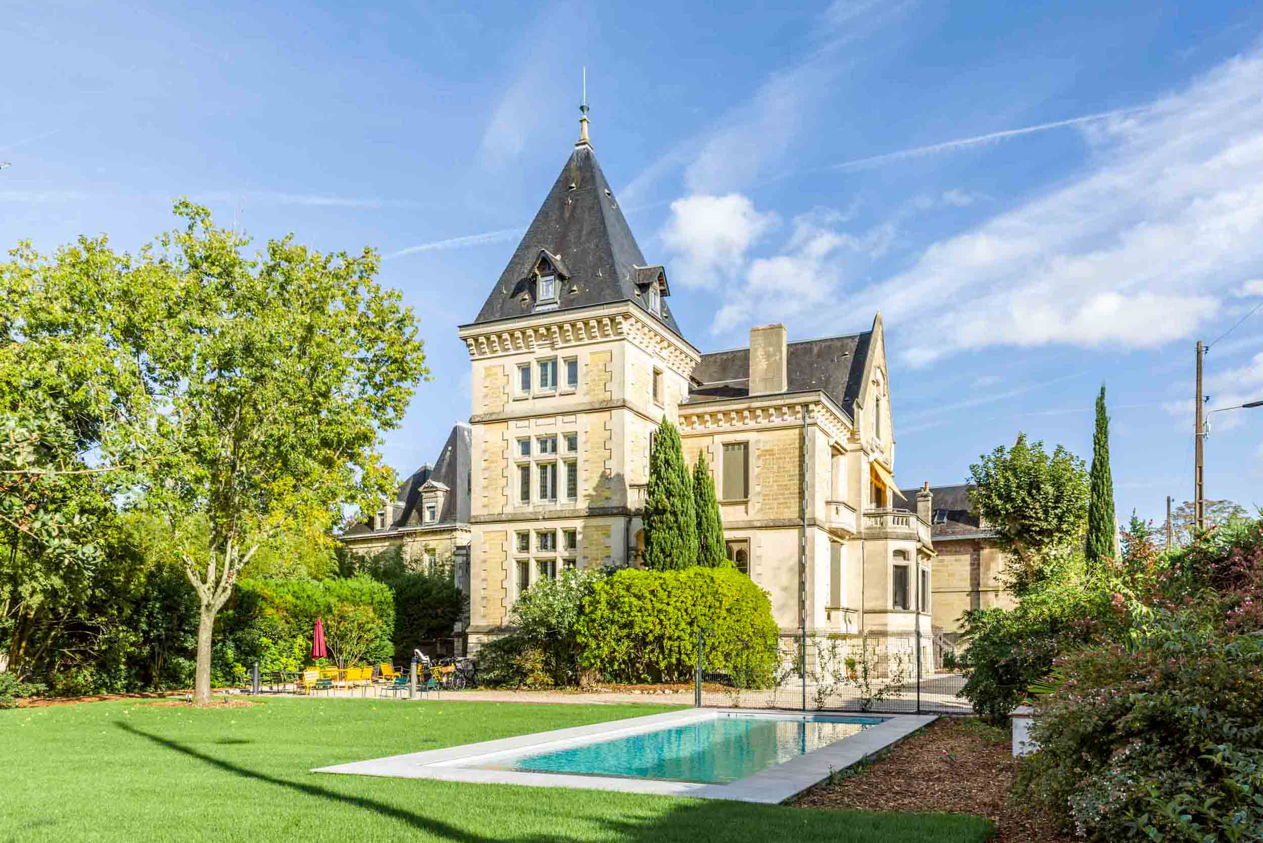 Une élégante demeure en pierre avec une tourelle pointue et des toits en ardoise pentus se dresse au milieu d'une végétation luxuriante sous un ciel bleu vif. Au premier plan, une piscine rectangulaire immaculée et une pelouse bien entretenue renforcent l'ambiance sereine. Des arbres et des buissons entourent la propriété, offrant intimité et charme, rappelant l'architecture bordelaise.